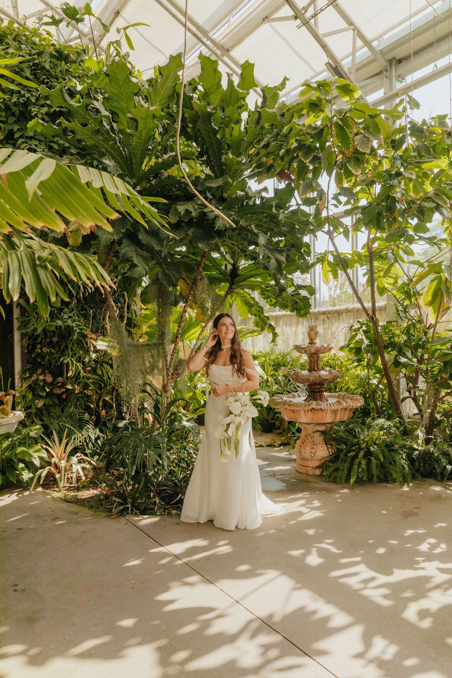 Bride in white off-shoulder gown holds bouquet beside stone fountain in lush tropical greenhouse.
