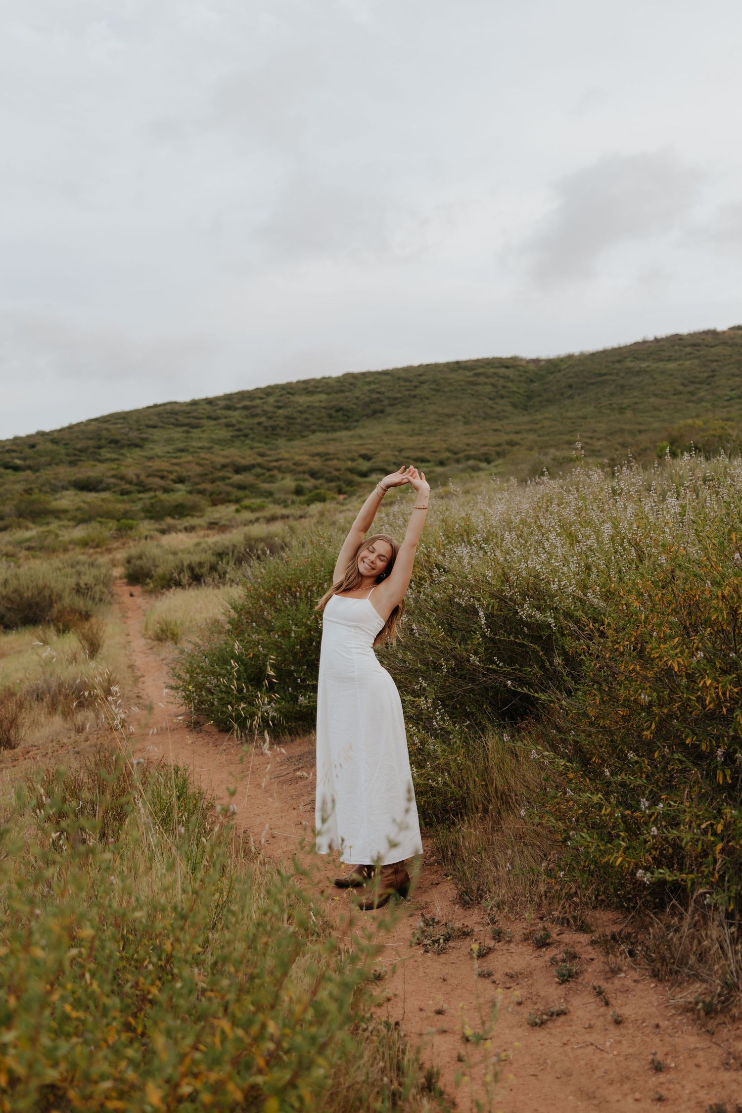 Woman in white dress stretching arms overhead on a scenic dirt trail surrounded by green hills.