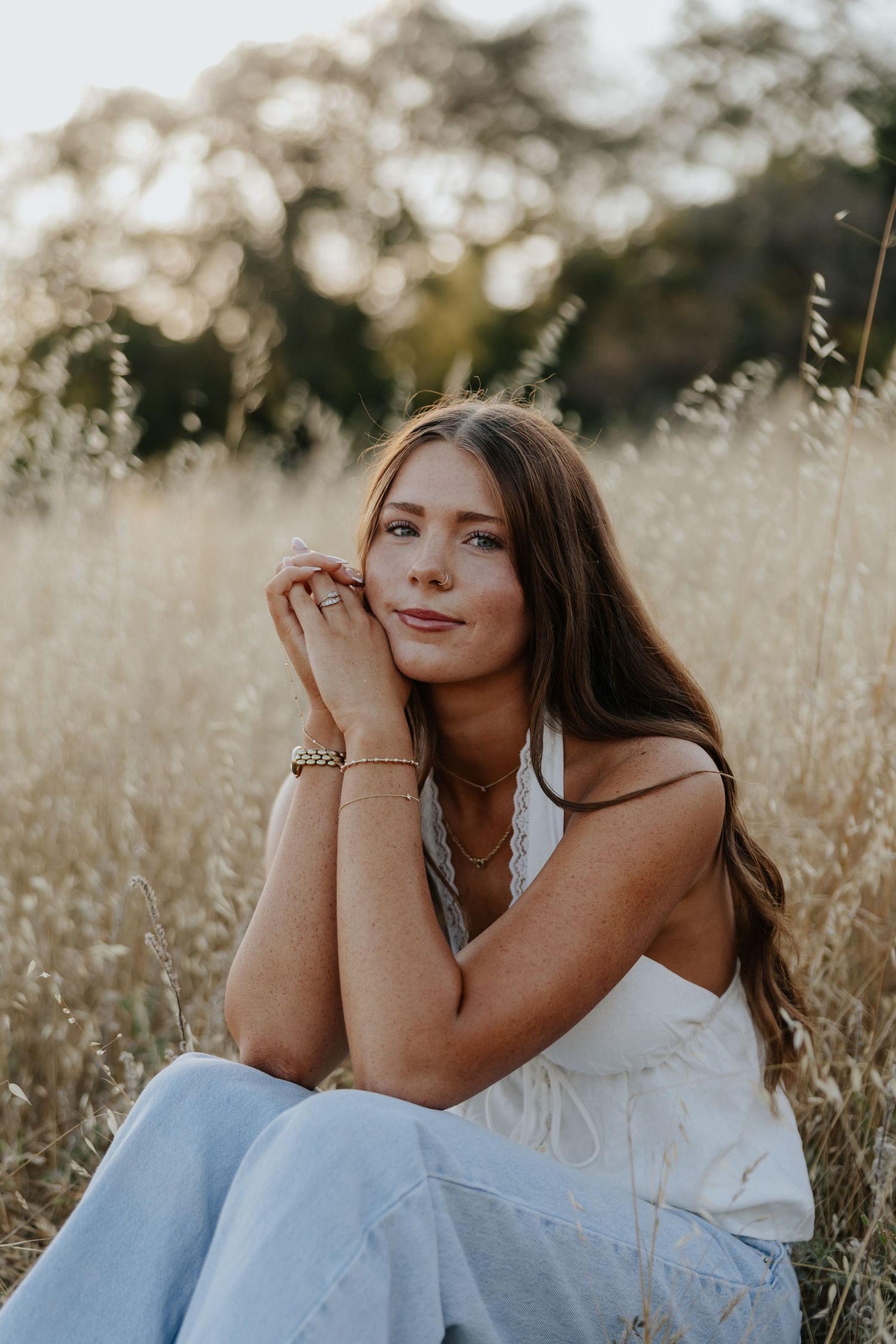 Young woman in white top and jeans sitting in tall golden grass field at sunset.
