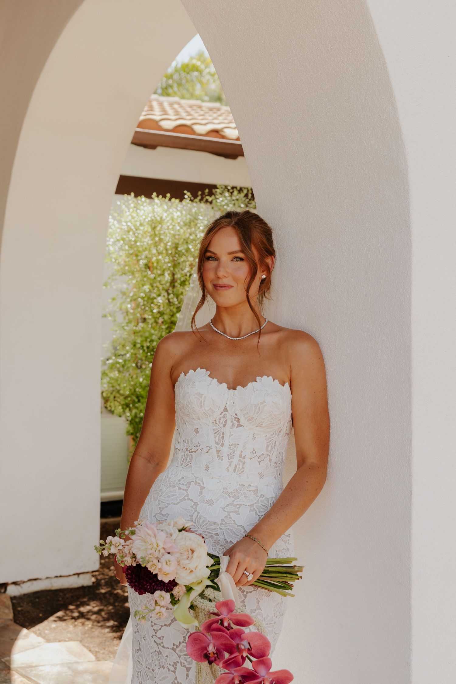 Beautiful bride in strapless lace wedding gown holding floral bouquet standing under white archway.