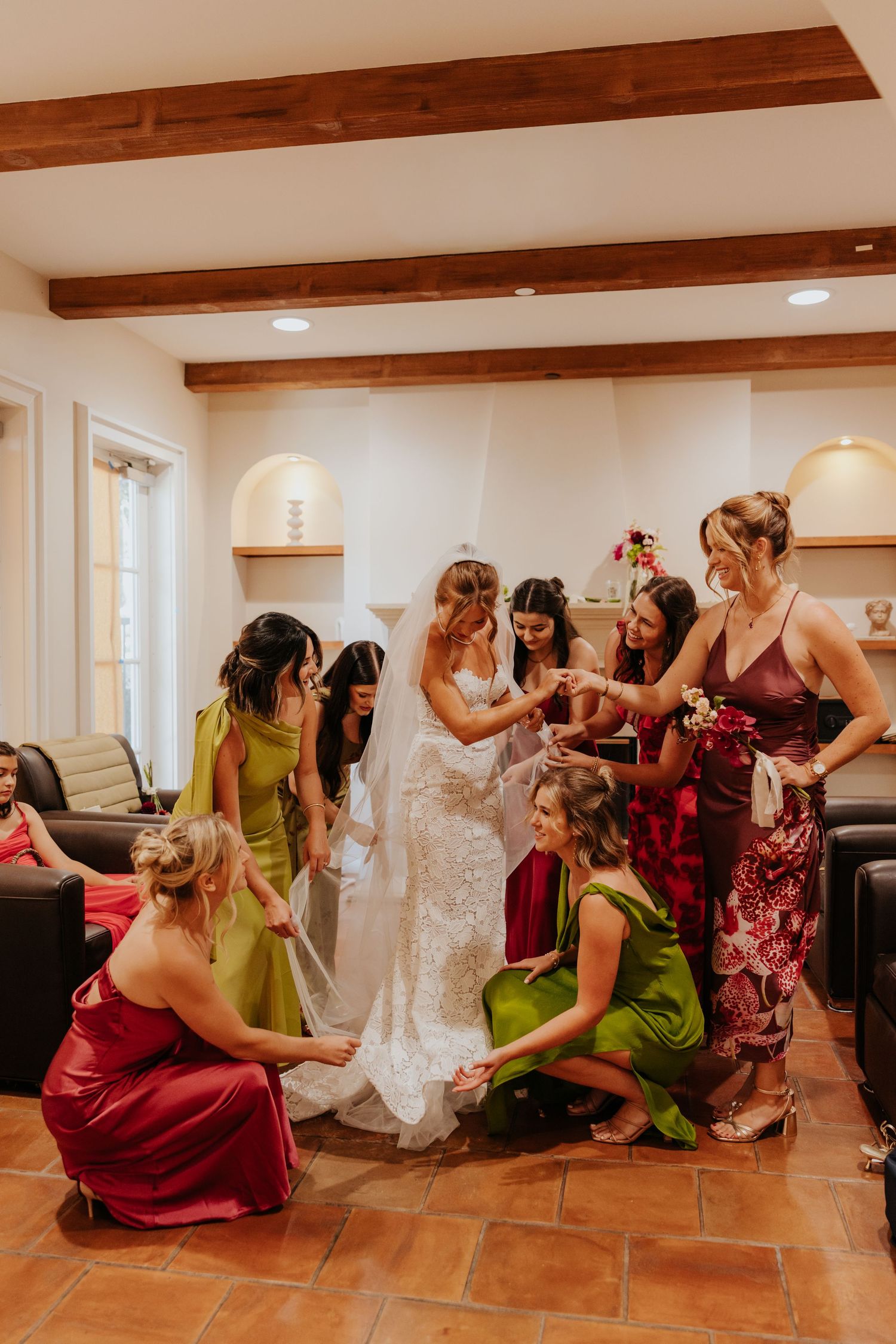Bride surrounded by bridesmaids in colorful dresses being helped with her lace wedding gown.