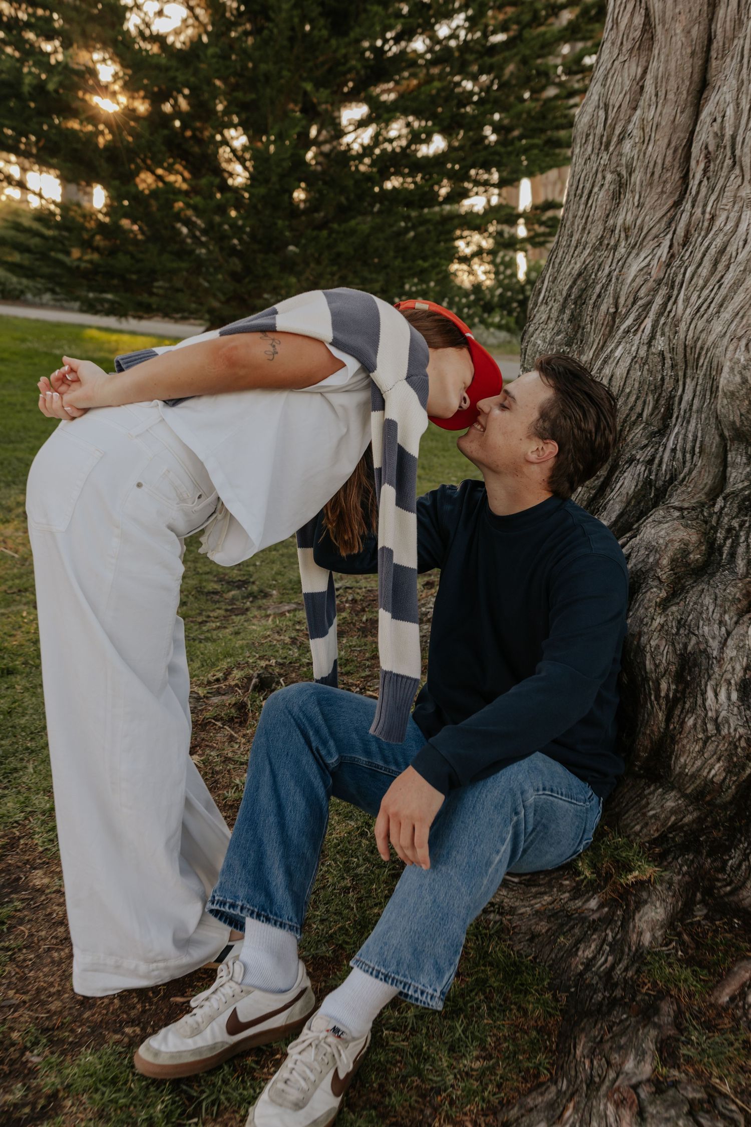 Couple sharing a kiss near a tree at sunset, woman in red cap bending down to kiss seated man.