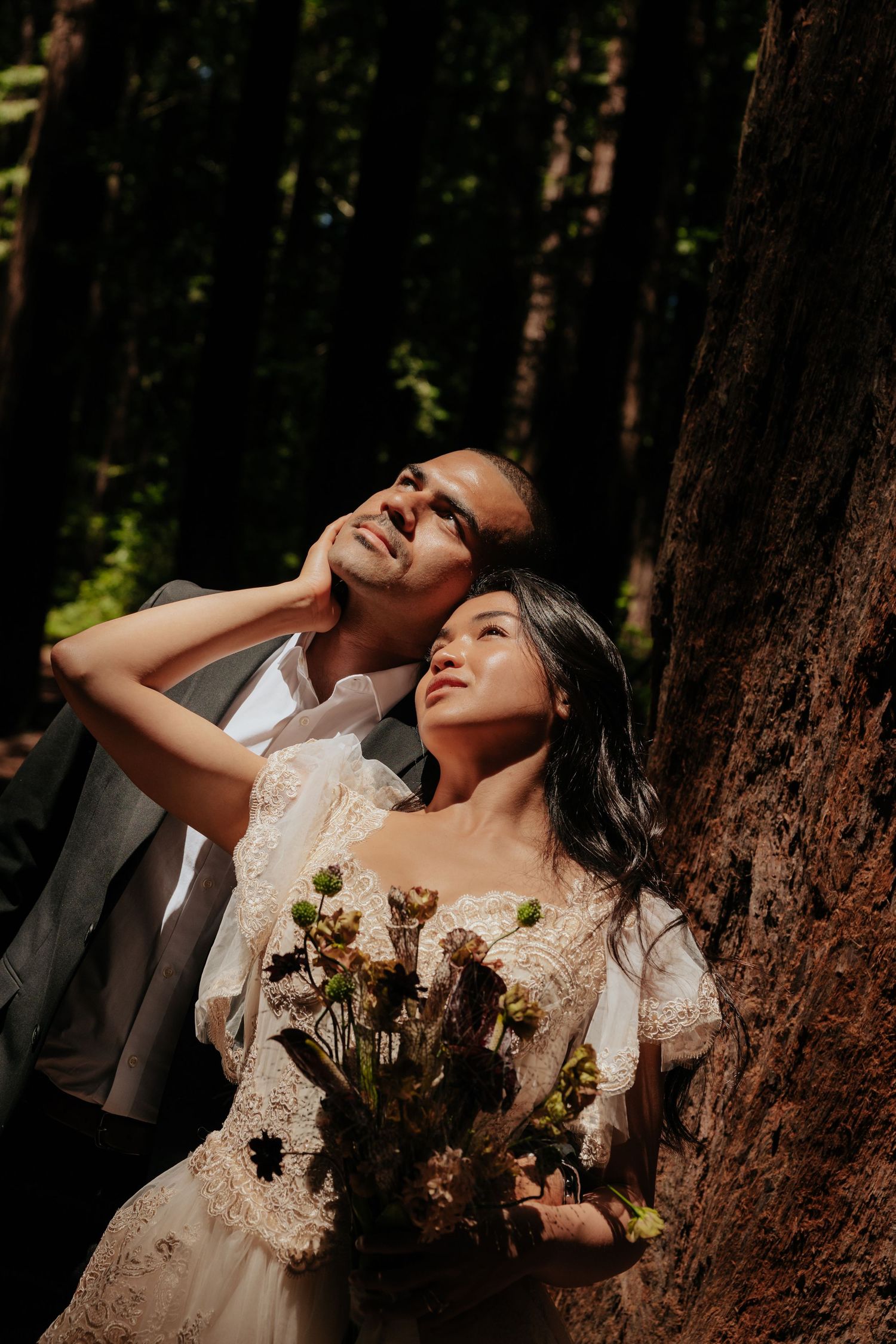 Couple in wedding attire embracing among tall redwood trees in dappled sunlight.
