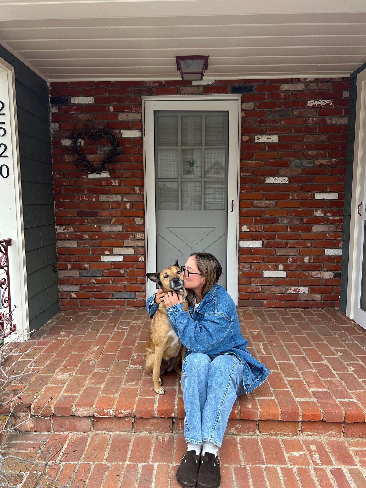 Young woman in denim jacket sitting on brick porch steps cuddling with a tan dog.