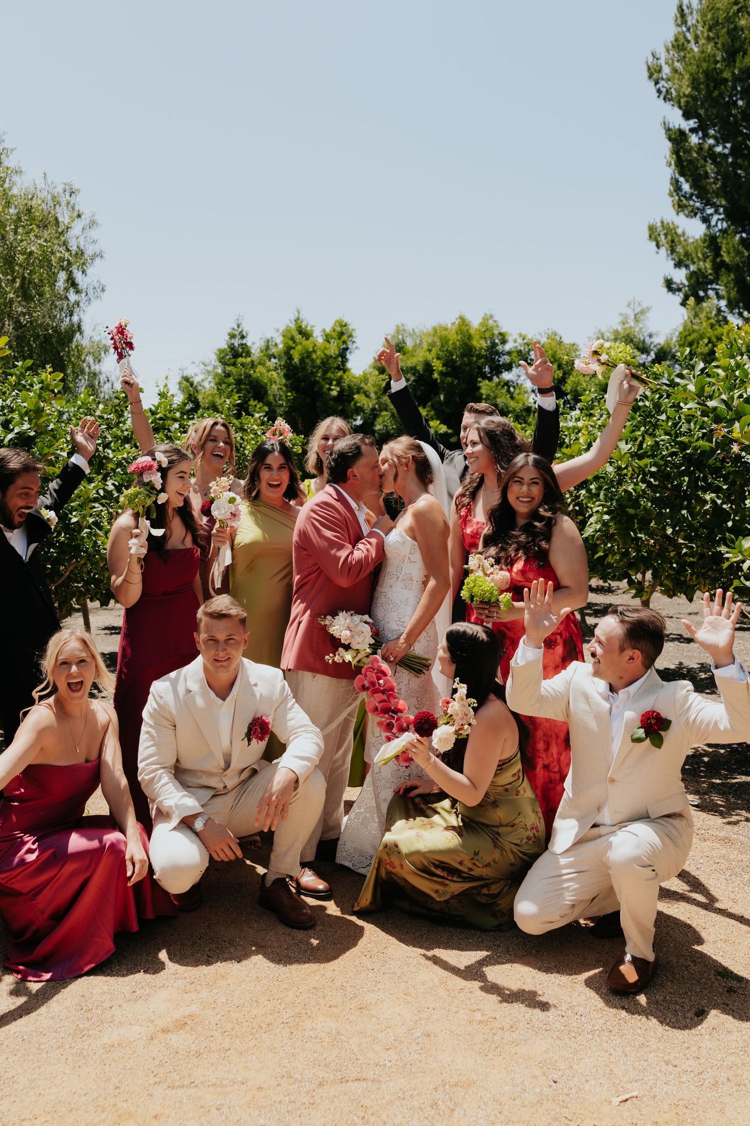 Joyful wedding party celebrating outdoors with bride and groom kissing surrounded by cheering guests.
