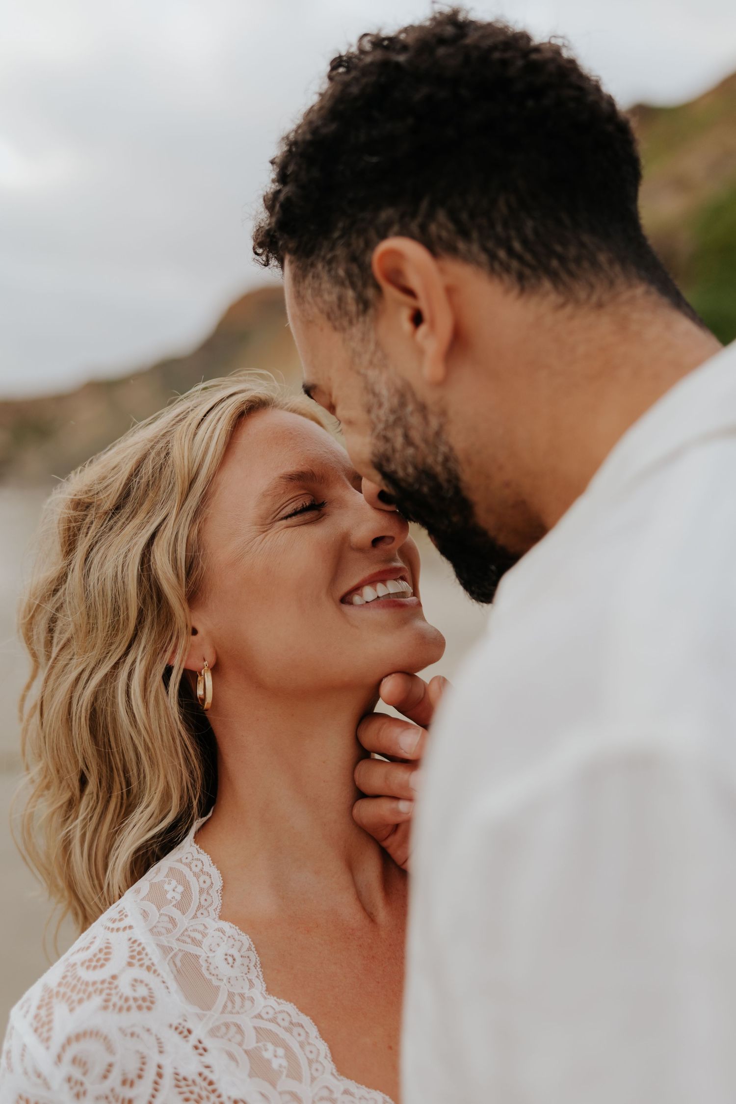 Smiling blonde woman in lace top gazes at curly-haired man gently holding her face outdoors.