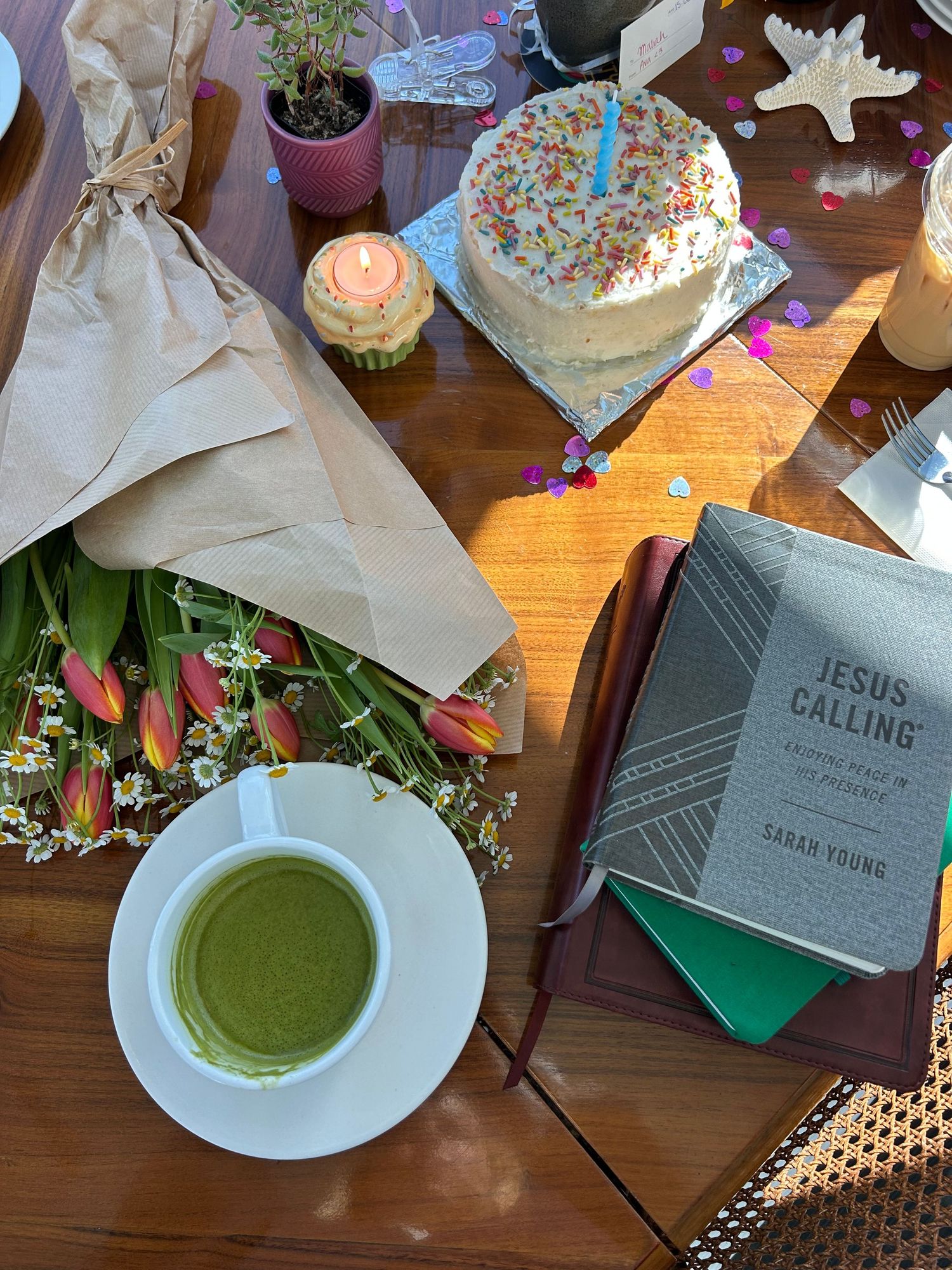 Birthday celebration table with matcha latte, tulips, Jesus Calling devotional book, and colorful sprinkle cake with candle.