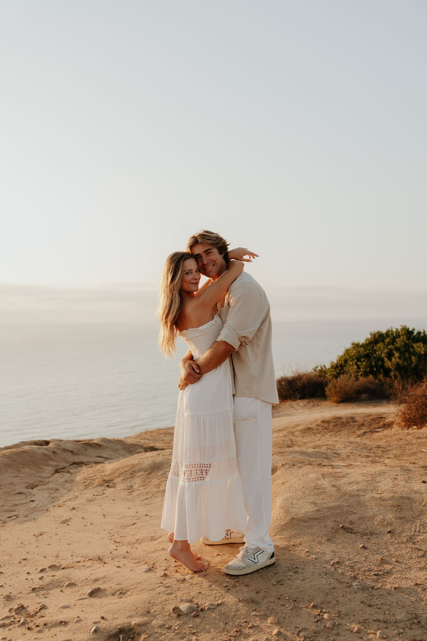 Couple embracing on sandy cliff at sunset, woman in white dress, man in white outfit, ocean backdrop.