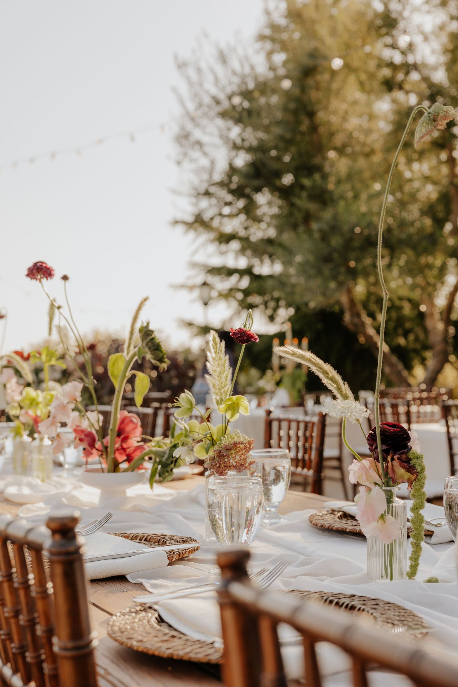 Elegant outdoor wedding reception table with wildflower centerpieces, woven chargers, and string lights.