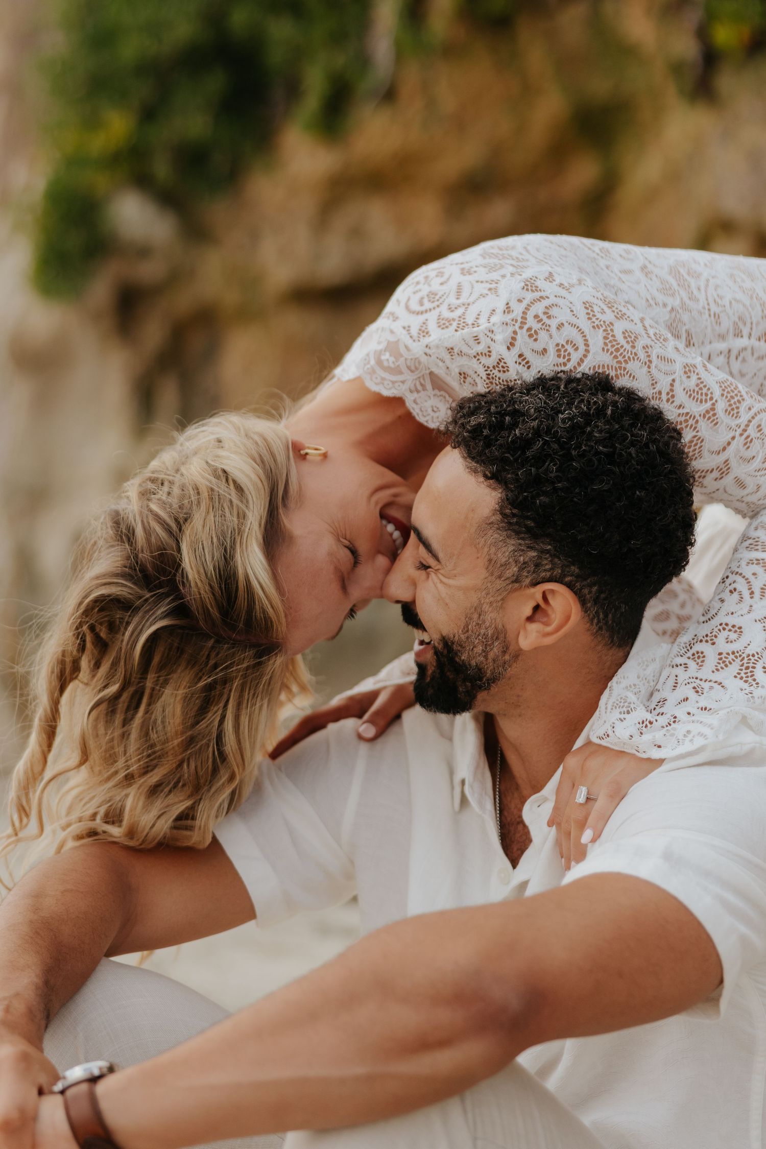 Joyful couple nearly kissing as groom lifts bride in white lace dress against rocky outdoor backdrop.