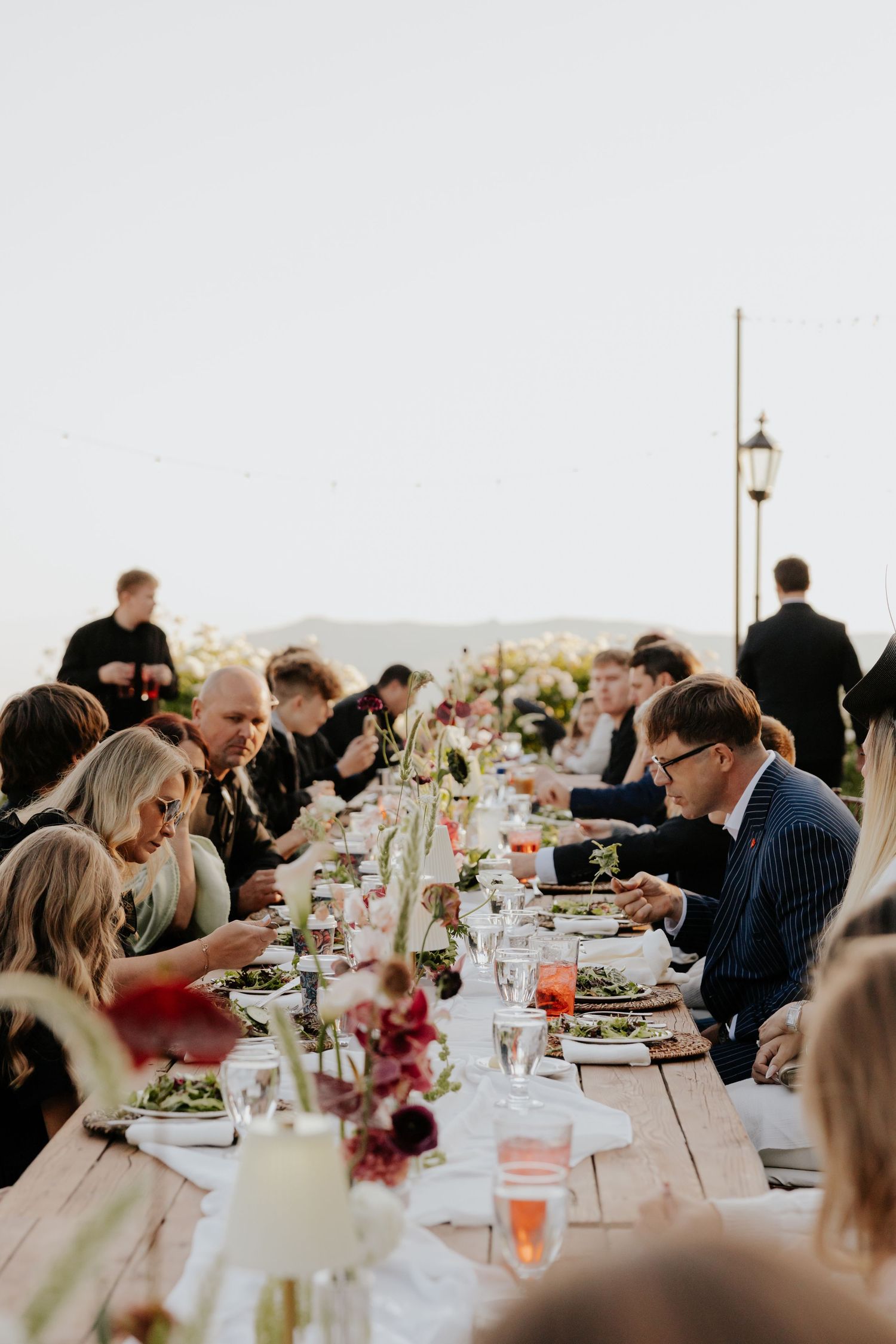 Guests dining at long rustic farm table adorned with floral centerpieces at outdoor wedding reception.