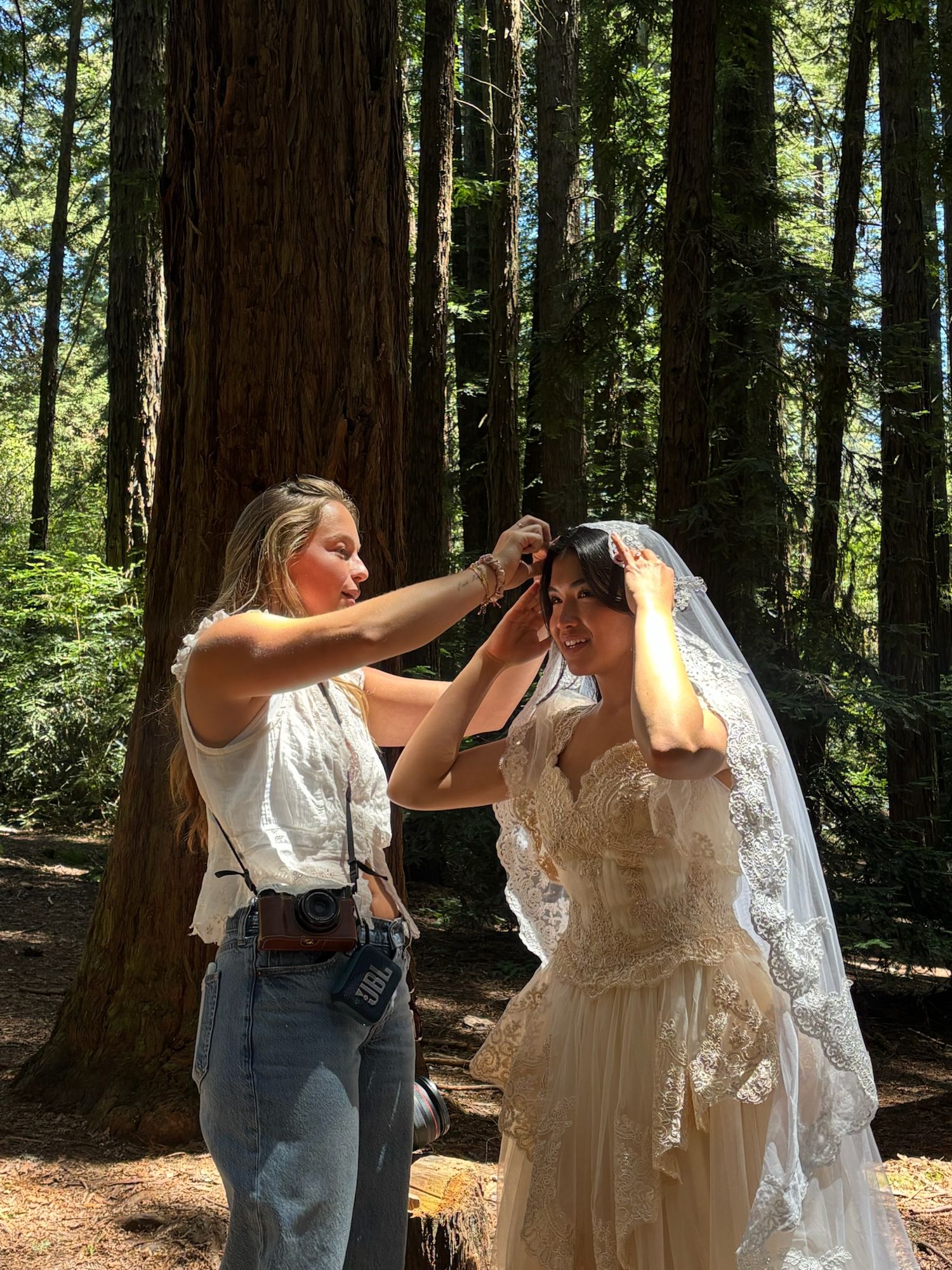 Blonde photographer adjusting veil on bride in lace dress among tall redwood trees.