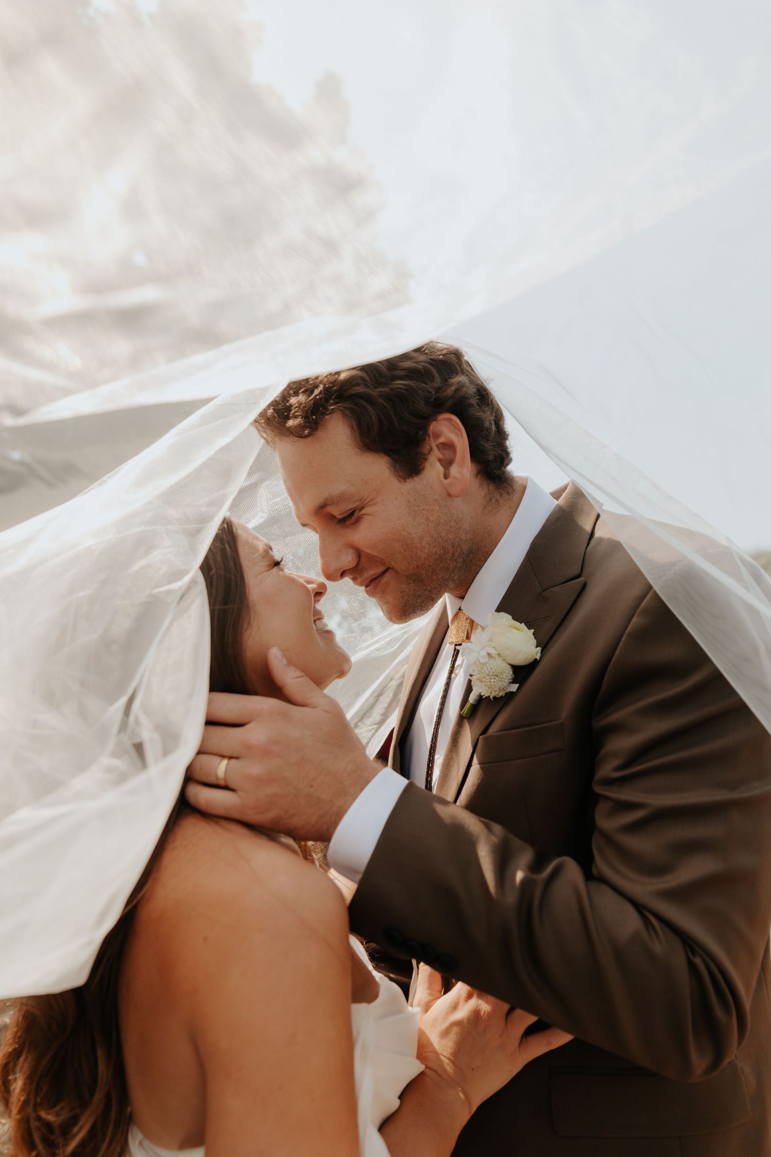 Bride and groom share romantic almost-kiss under flowing veil in warm golden sunlight.