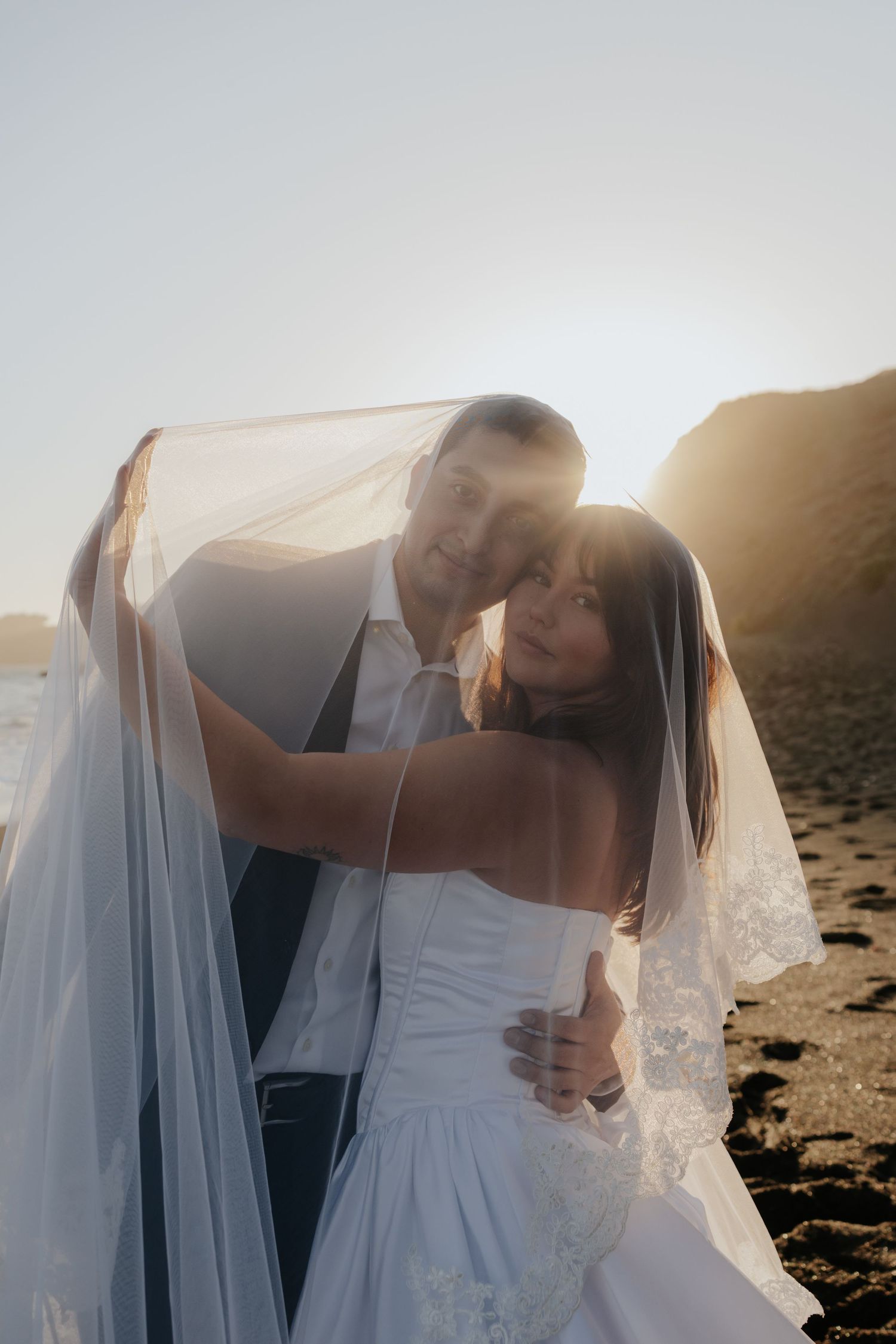 Bride and groom embrace under a lace veil with golden sunset light on a rocky beach.