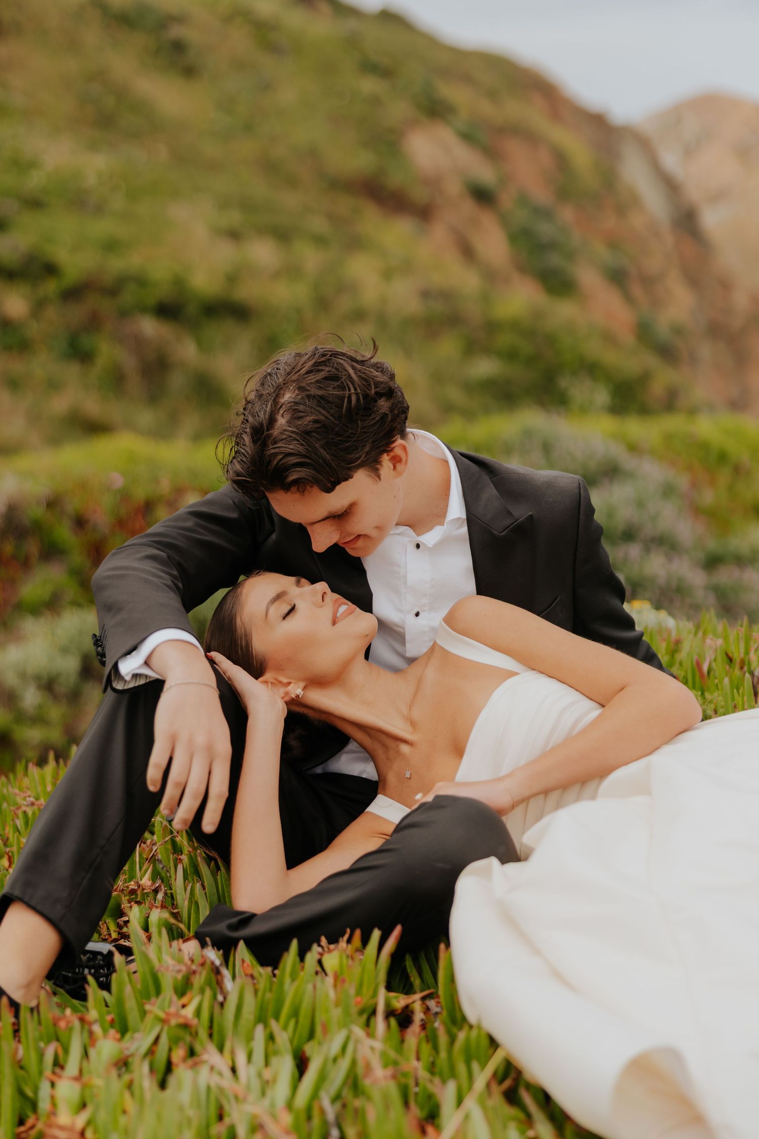 Bride and groom share a romantic moment lying in green coastal meadow grass with mountain backdrop.