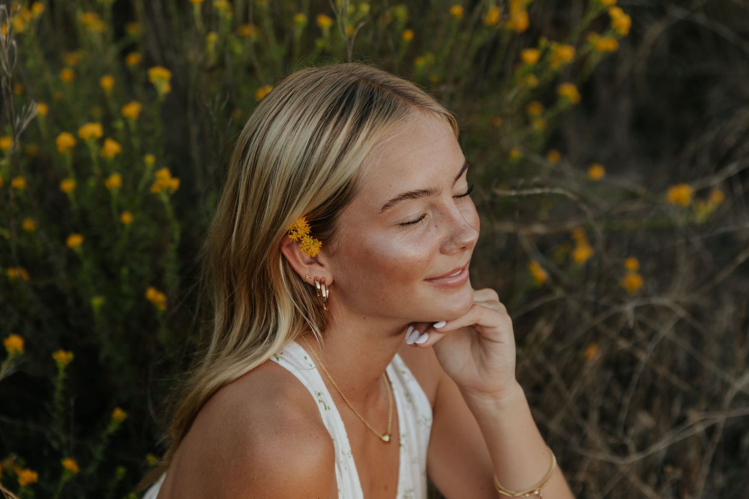 Close-up of smiling blonde woman with yellow flower in hair sitting among wildflowers in warm sunlight.