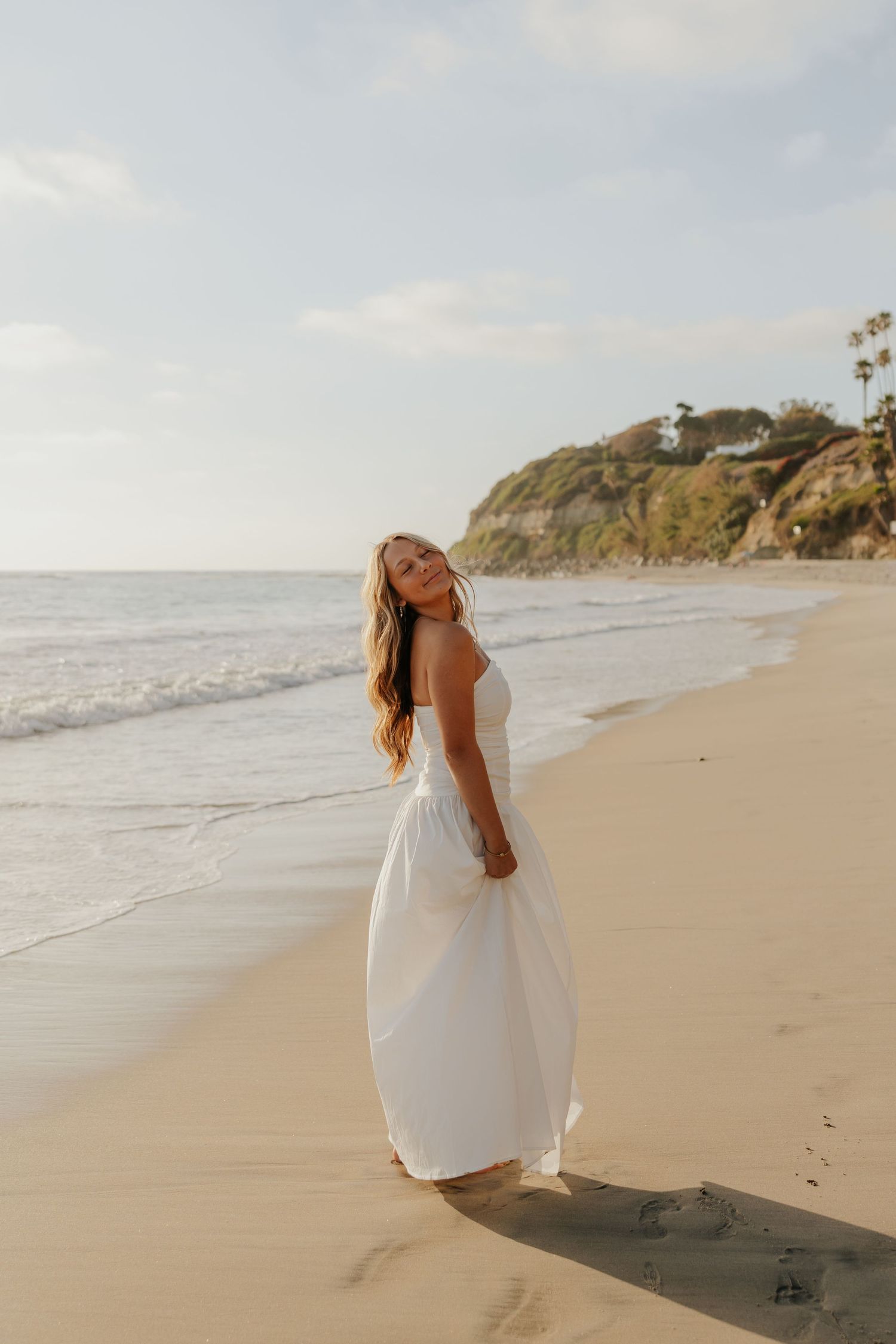 Blonde woman in white maxi dress standing on sandy beach at golden hour with ocean waves and cliffside in background.