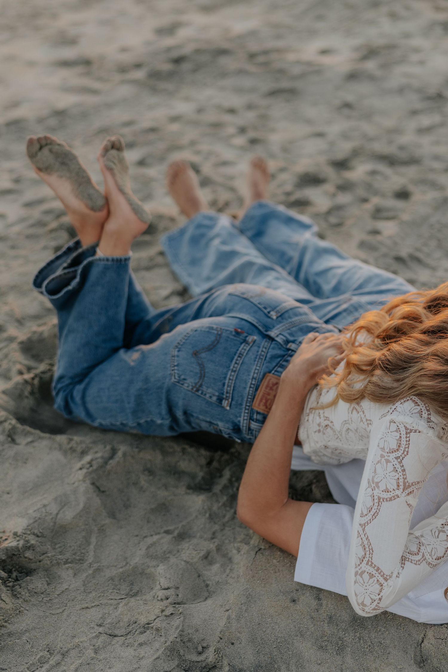 Couple lying together in sand on beach, wearing jeans and white tops.