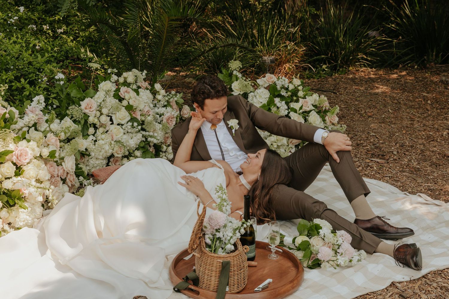 Bride and groom share intimate moment lying on picnic blanket surrounded by white floral arrangements.