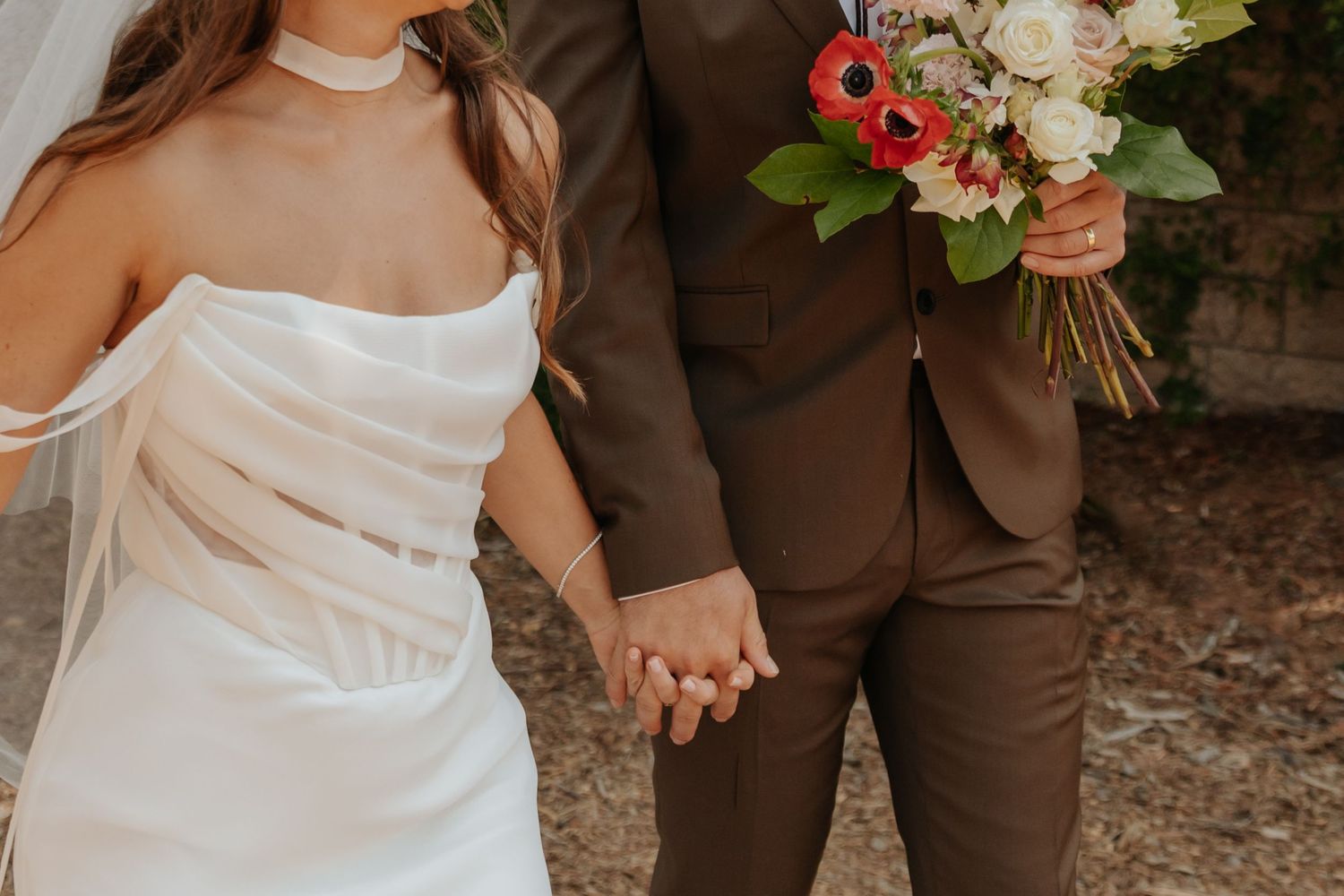 Bride and groom hold hands, she wears white strapless gown, he wears brown suit with colorful bouquet.