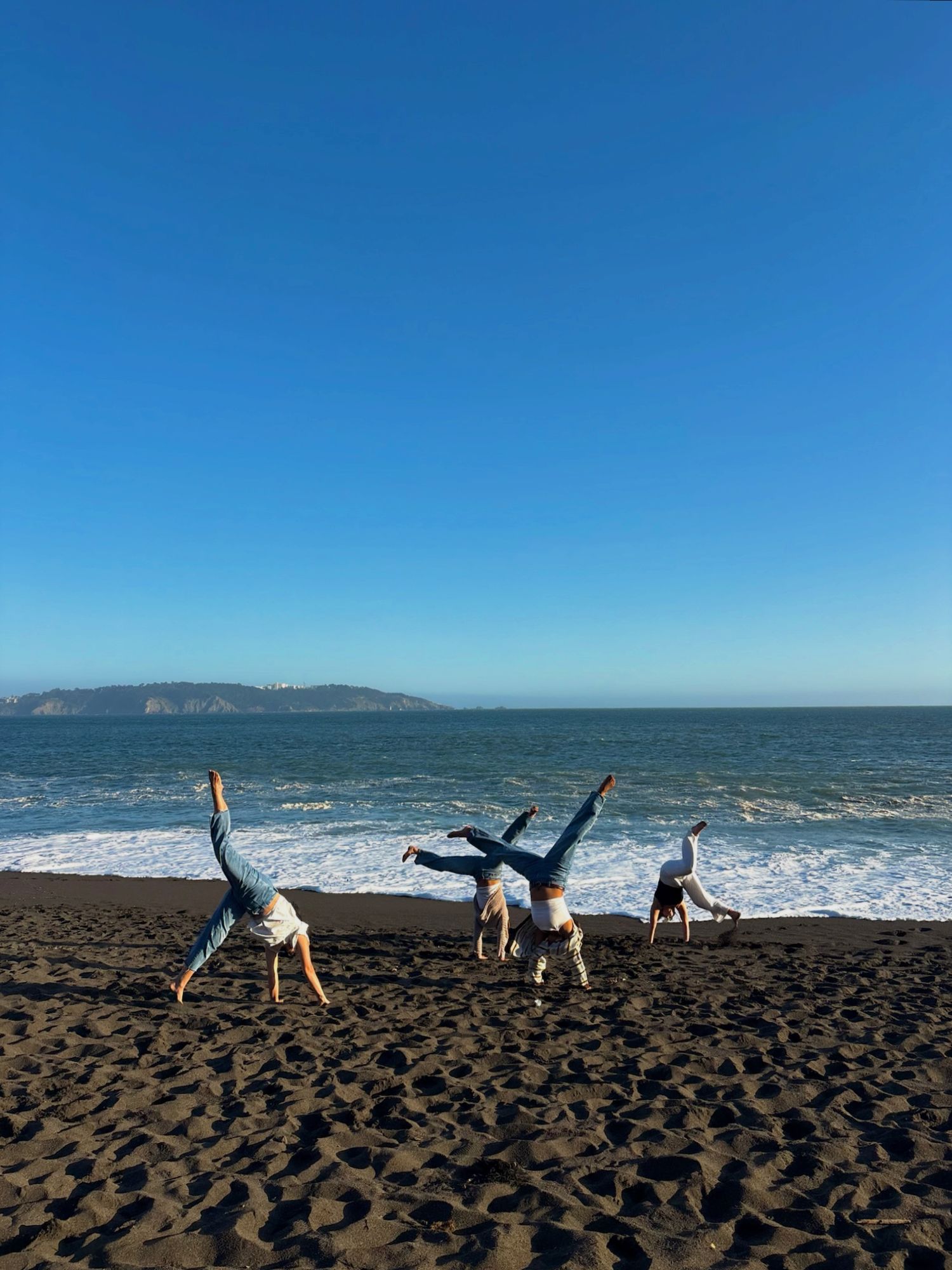 Four people doing simultaneous cartwheels on dark sand beach with blue ocean backdrop.
