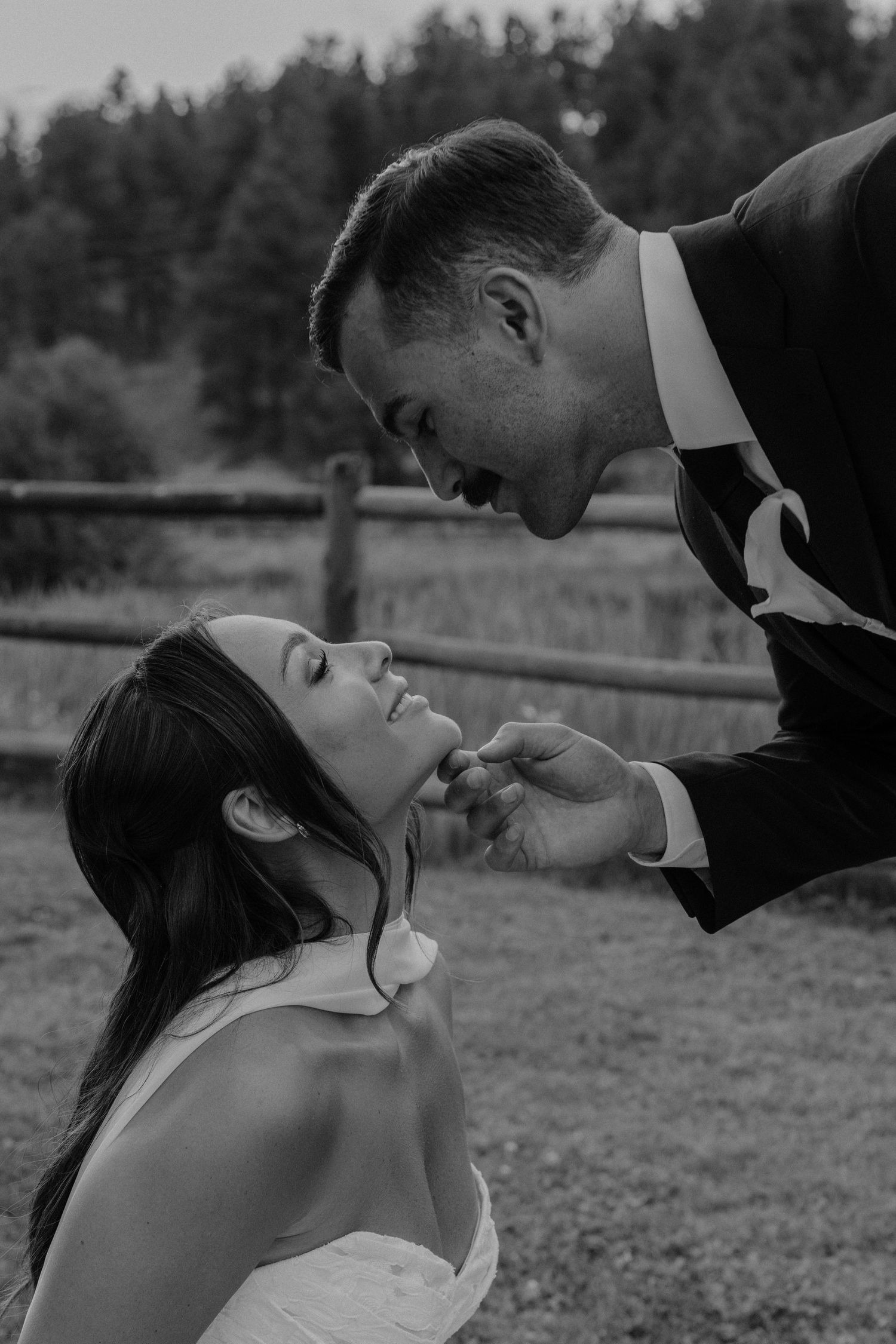 Black and white photo of a groom tenderly lifting bride's chin, sharing an intimate moment outdoors near a wooden fence.