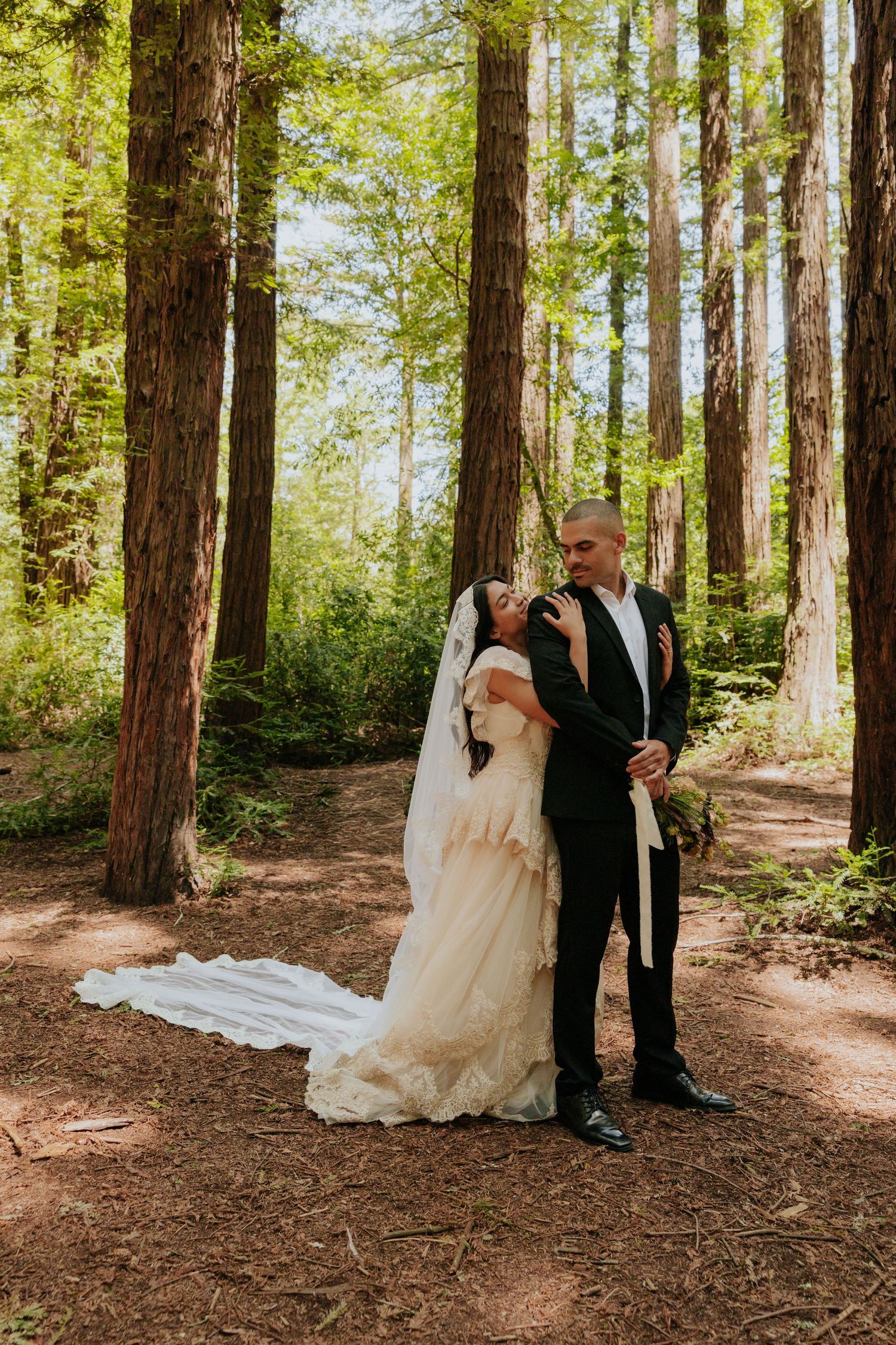 Bride and groom standing among tall redwood trees, bride in lace gown with long veil trailing on ground.