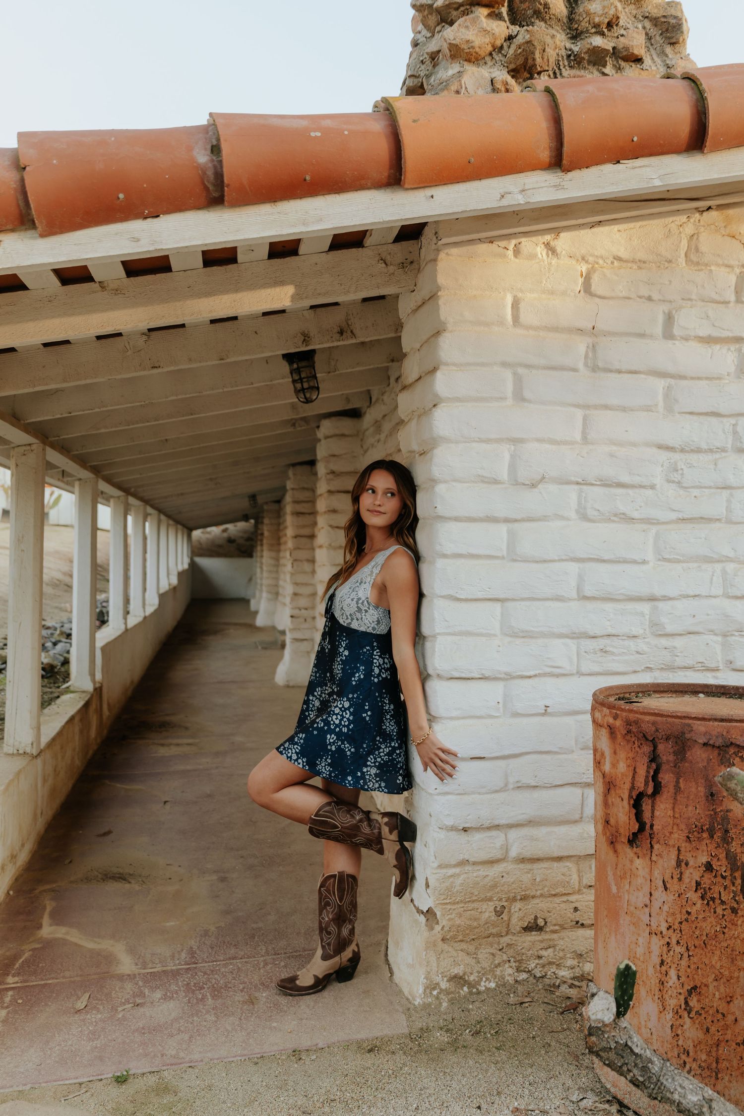 Brunette woman in floral skirt and cowboy boots leaning against white brick wall of rustic covered walkway.
