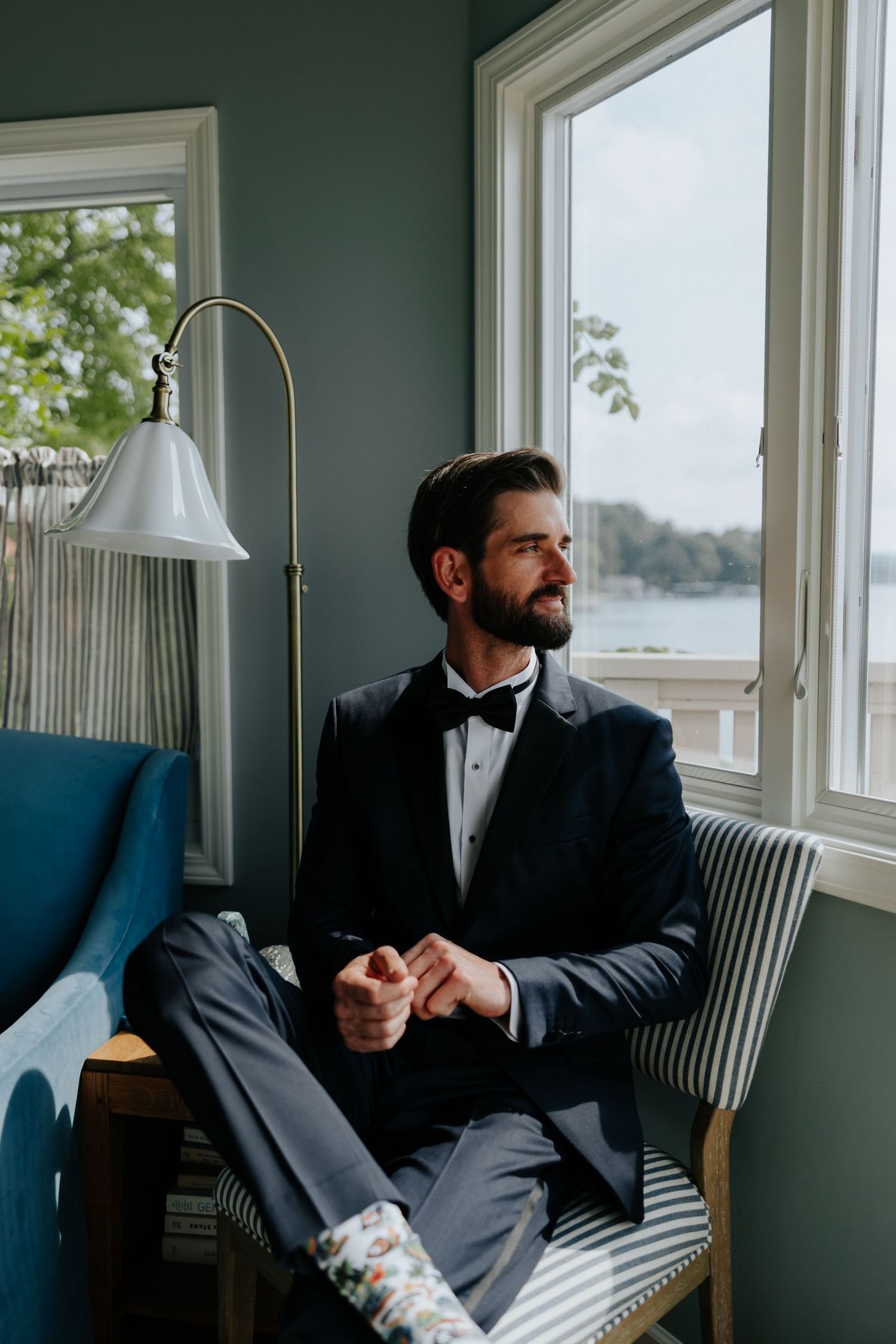 A dapper groom in a navy tuxedo and bow tie sits by a window, gazing outside on his wedding day.