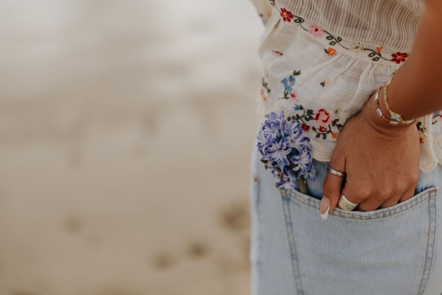 Close-up of woman in floral top and jeans with hand in pocket holding blue flowers on beach.