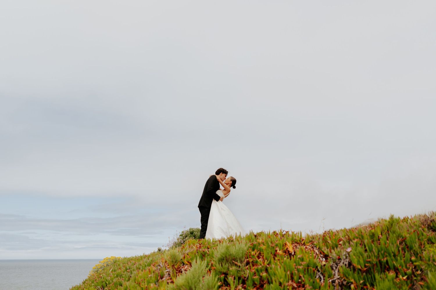 Bride and groom share a romantic dip kiss on a coastal hillside with ocean views.