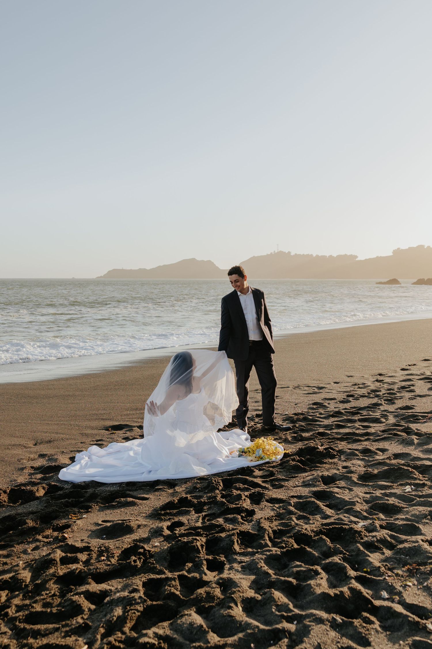 Bride kneels on sandy beach in veil while groom stands beside her at golden hour sunset.