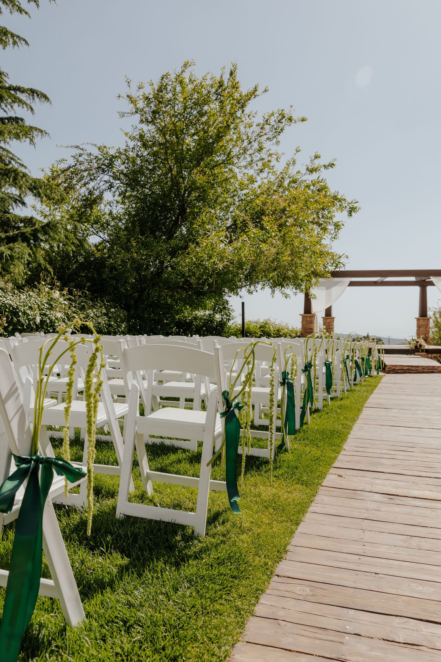 Outdoor wedding ceremony setup with white folding chairs, green ribbon accents, and wooden aisle.