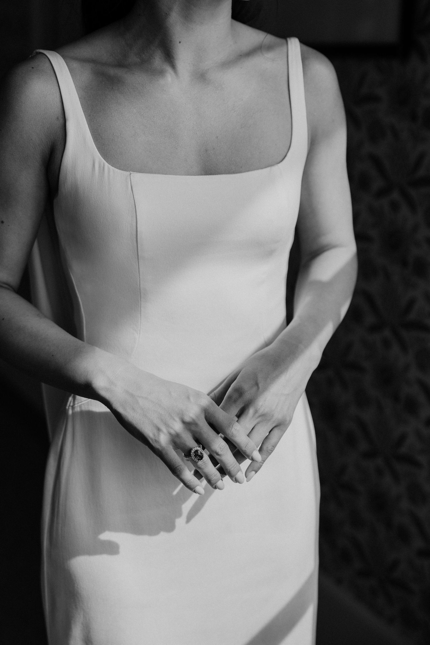 Black and white photo of a bride in a sleek white gown, hands clasped showing a statement ring.