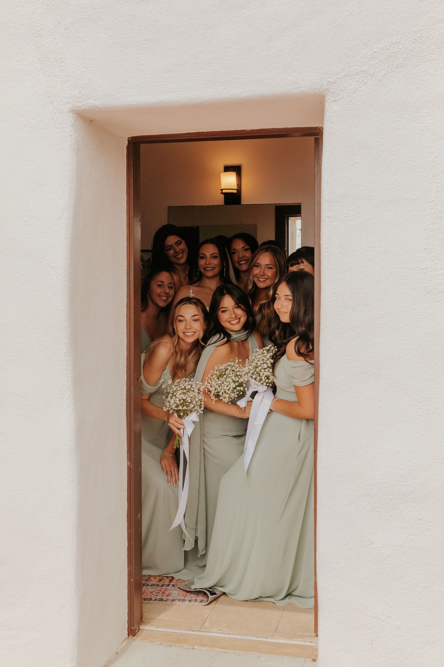 Bridesmaids in sage green gowns holding baby's breath bouquets gathered in doorway before wedding ceremony.