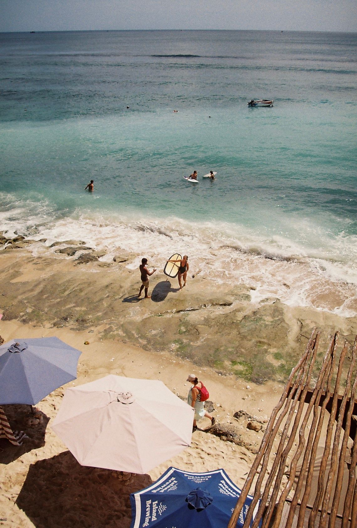 Aerial view of a tropical beach with umbrellas, surfers, and swimmers enjoying crystal-clear turquoise ocean waters.