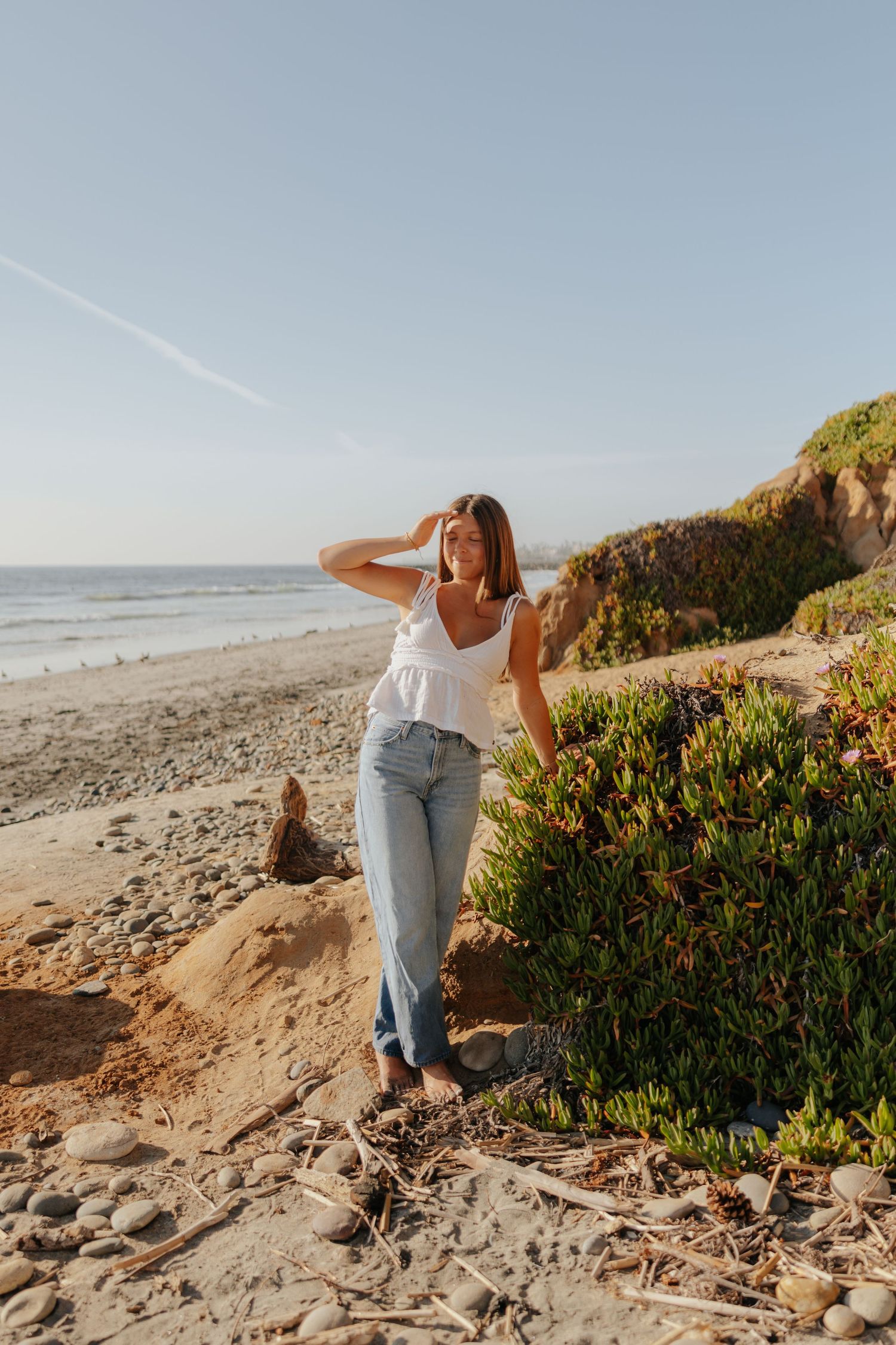 Young woman in white top and jeans poses on sandy beach with ocean and cliffs in background.