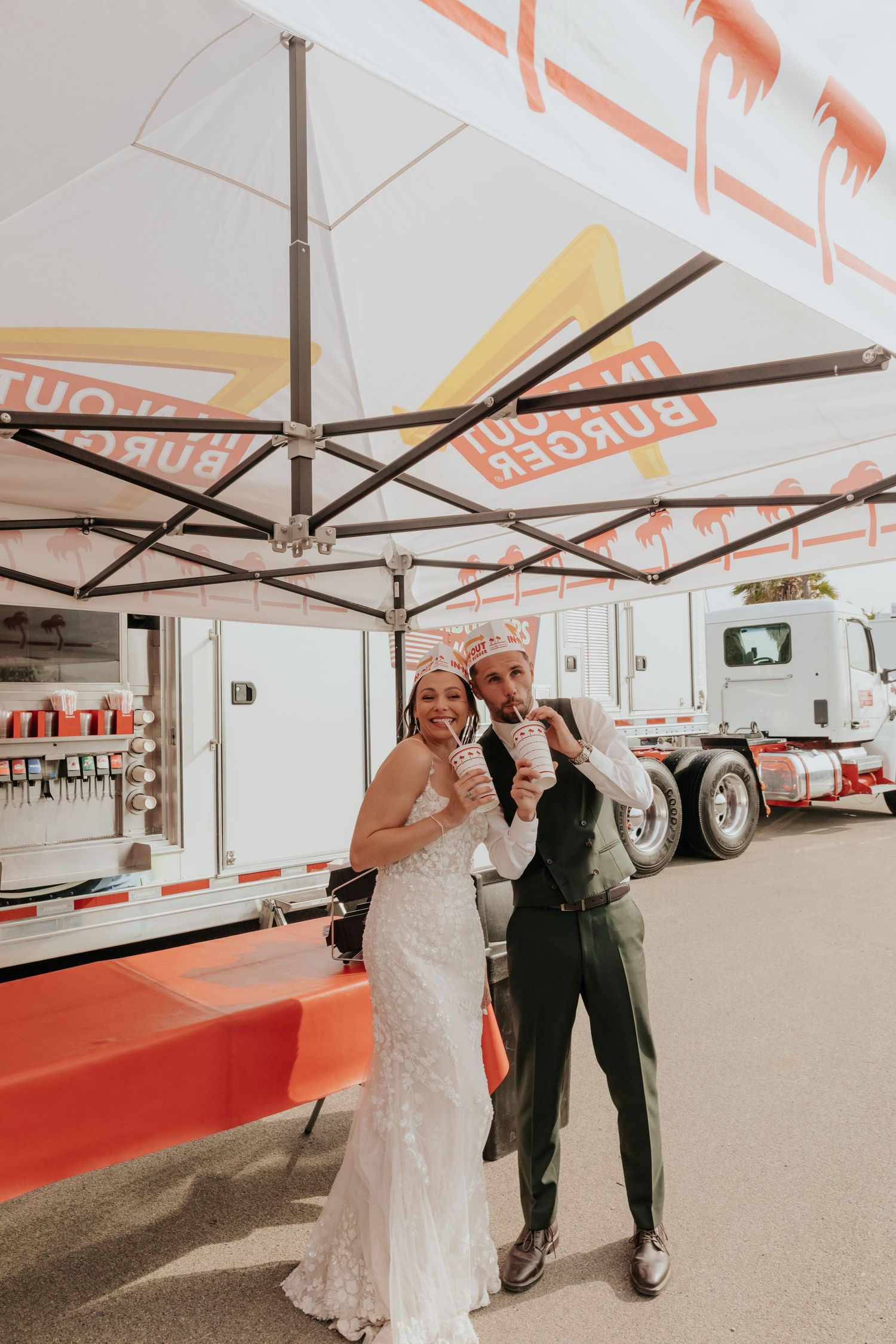 Smiling bride and groom in wedding attire enjoy milkshakes together at outdoor burger food truck reception.