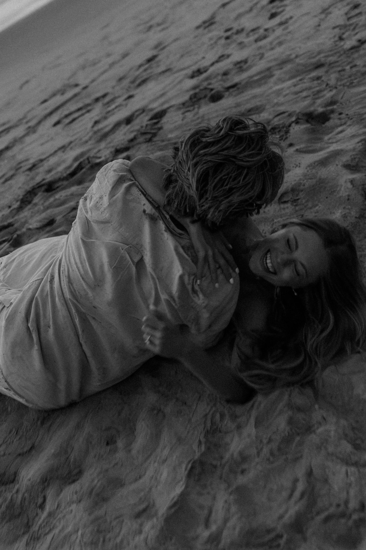 Black and white photo of a laughing woman lying on sandy beach with a child hugging her, captured in a joyful moment.