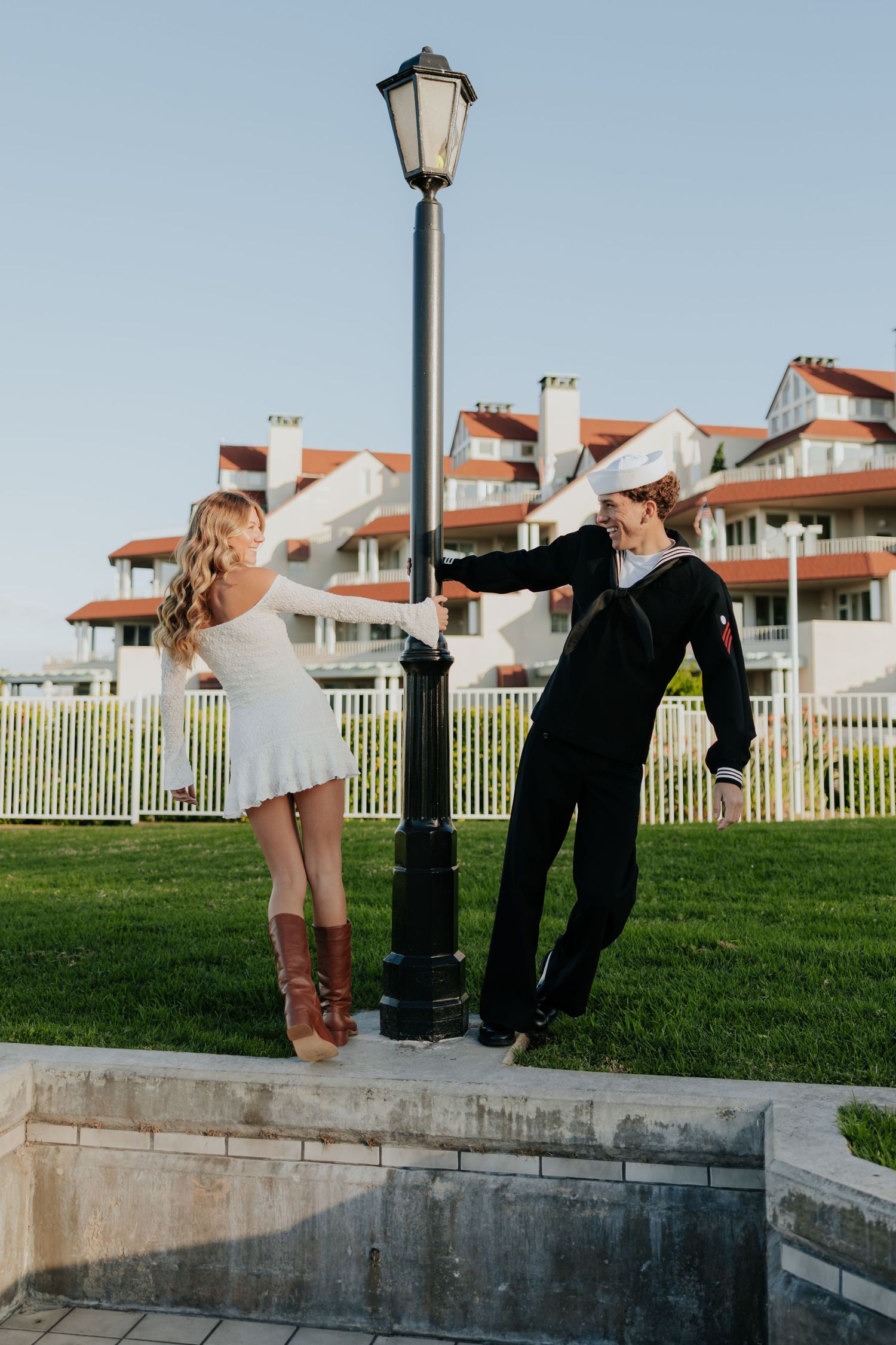 Blonde woman in white mini dress and sailor in uniform lean on lamppost on green lawn.