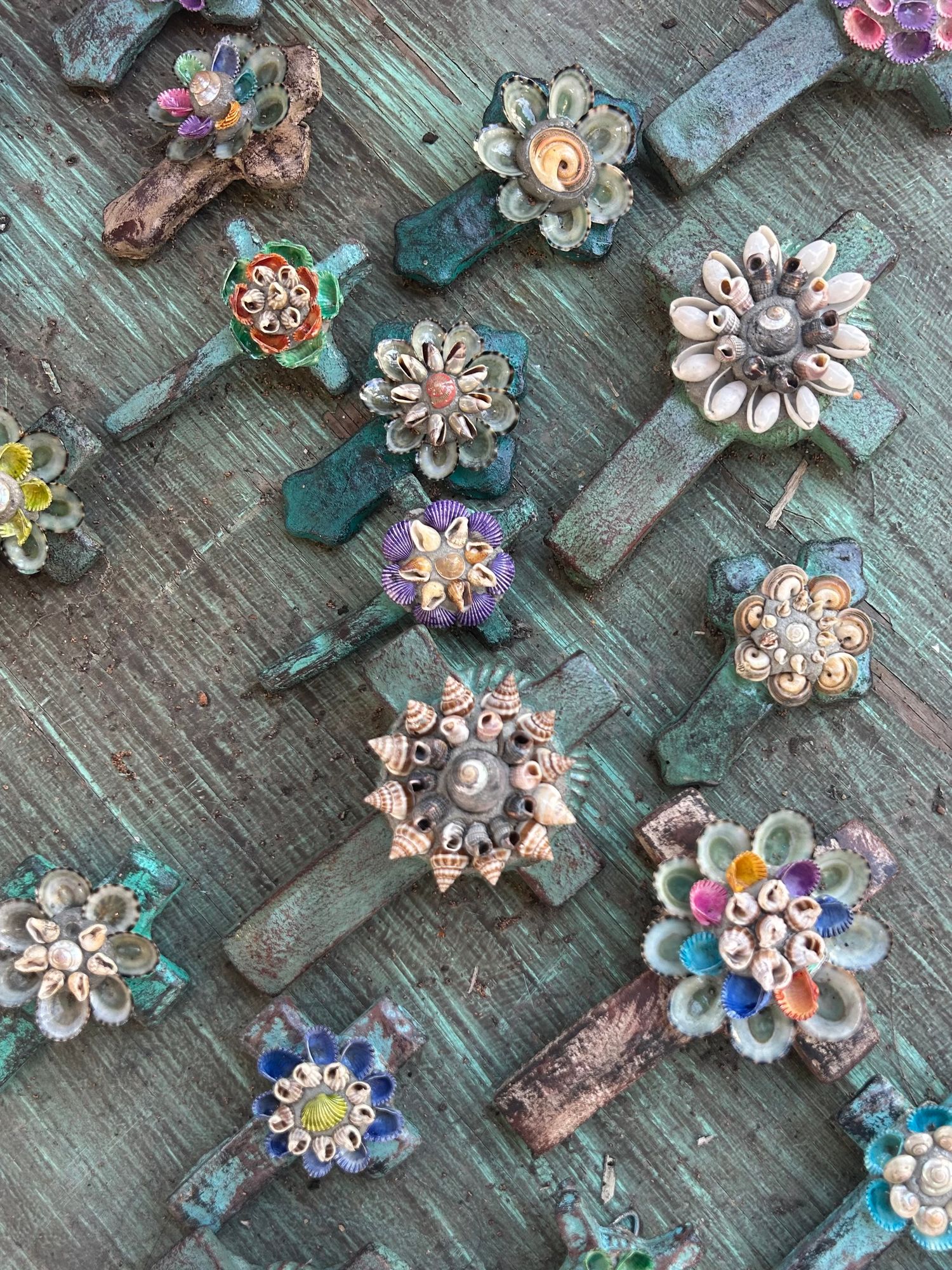 Handmade decorative crosses adorned with colorful shells and flowers on rustic teal wood.