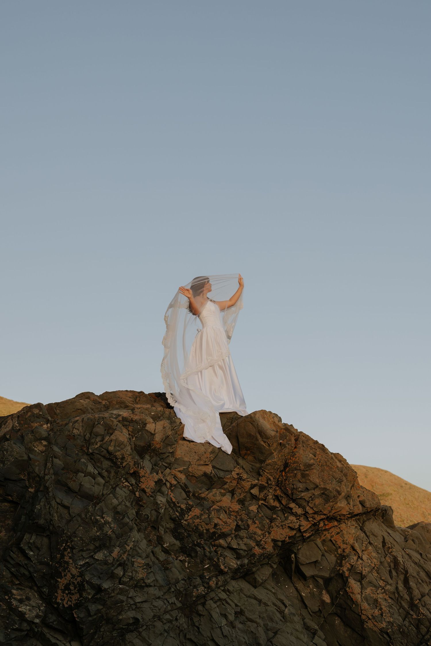 Bride in flowing white gown stands atop rocky cliffs holding her veil against blue sky.