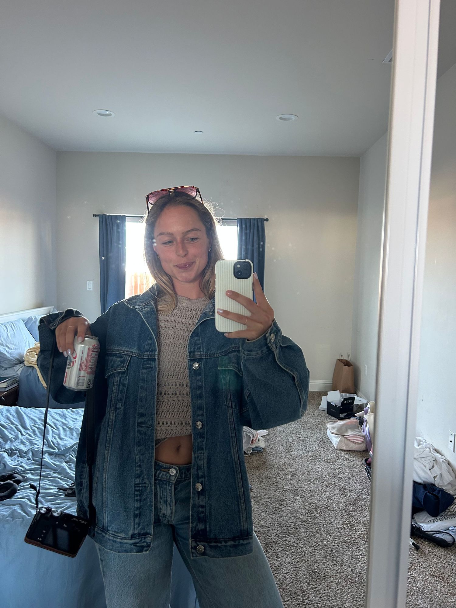Young woman in denim jacket taking mirror selfie holding a can in bedroom.