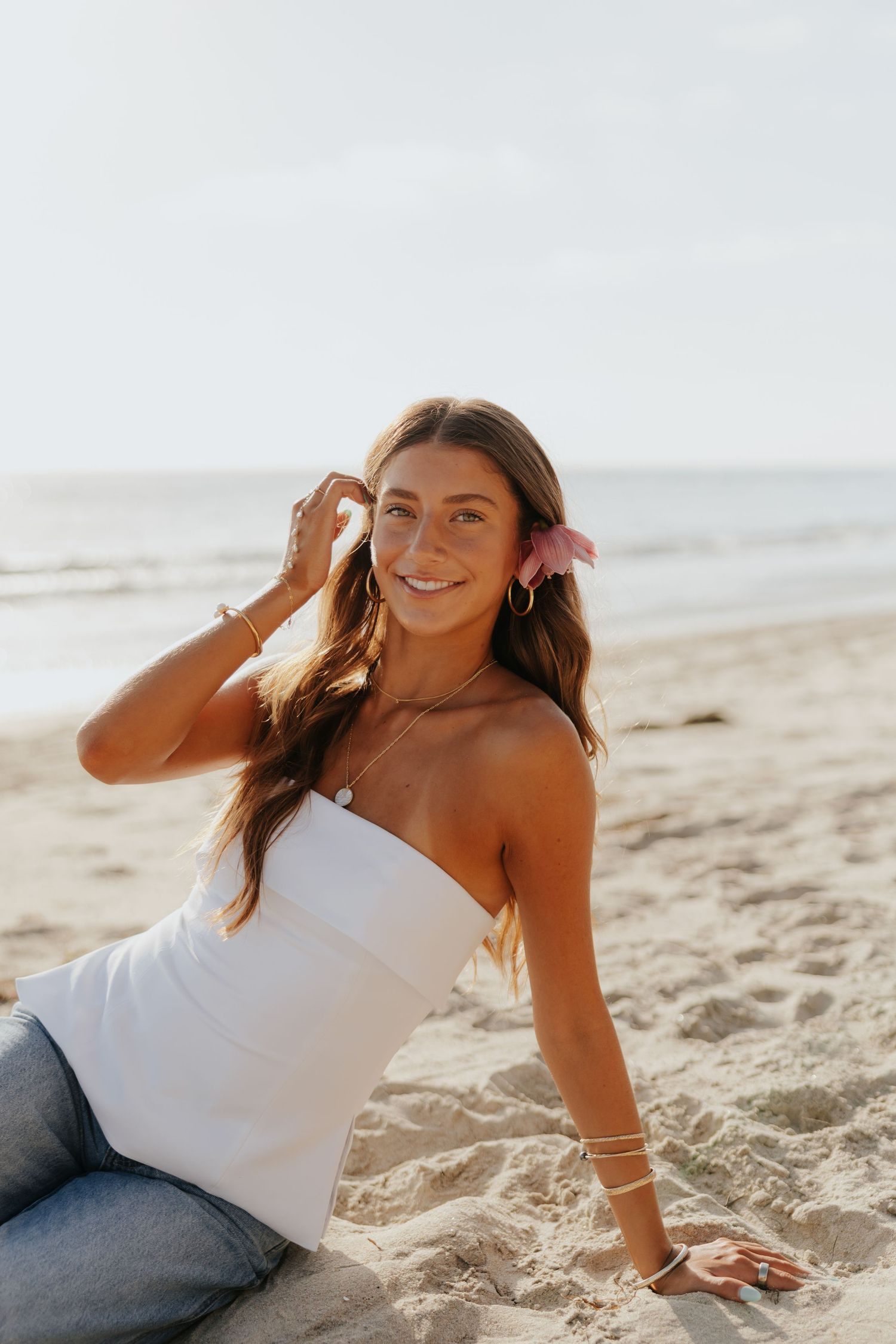 Smiling young woman with pink flower in hair sits on sandy beach in white top and jeans at sunset.