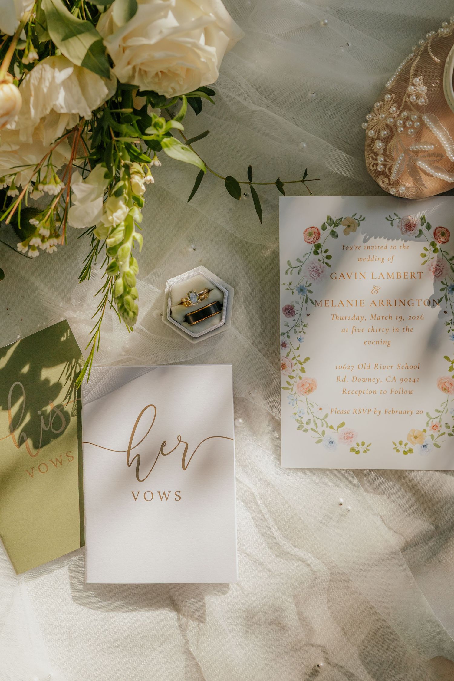 Wedding flat lay featuring floral invitation, vow booklet, ring box, and white roses on sheer fabric.