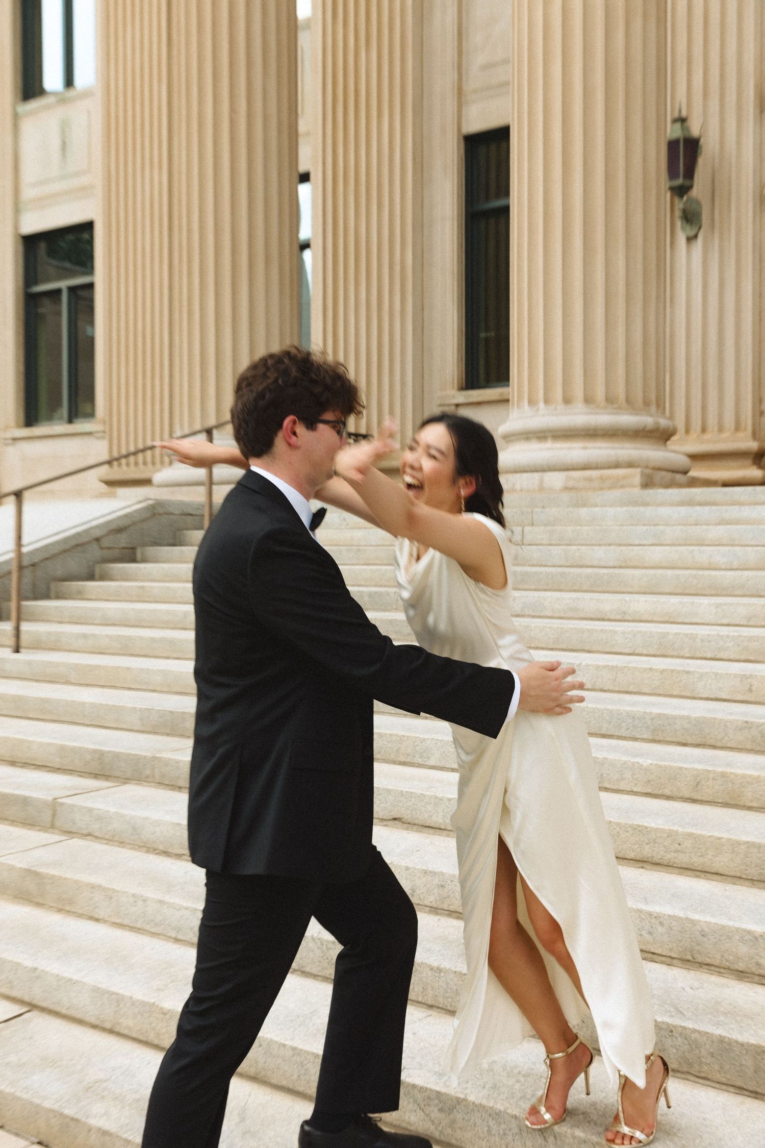 Laughing bride in off-shoulder satin dress dancing with tuxedo-clad groom on grand courthouse steps.