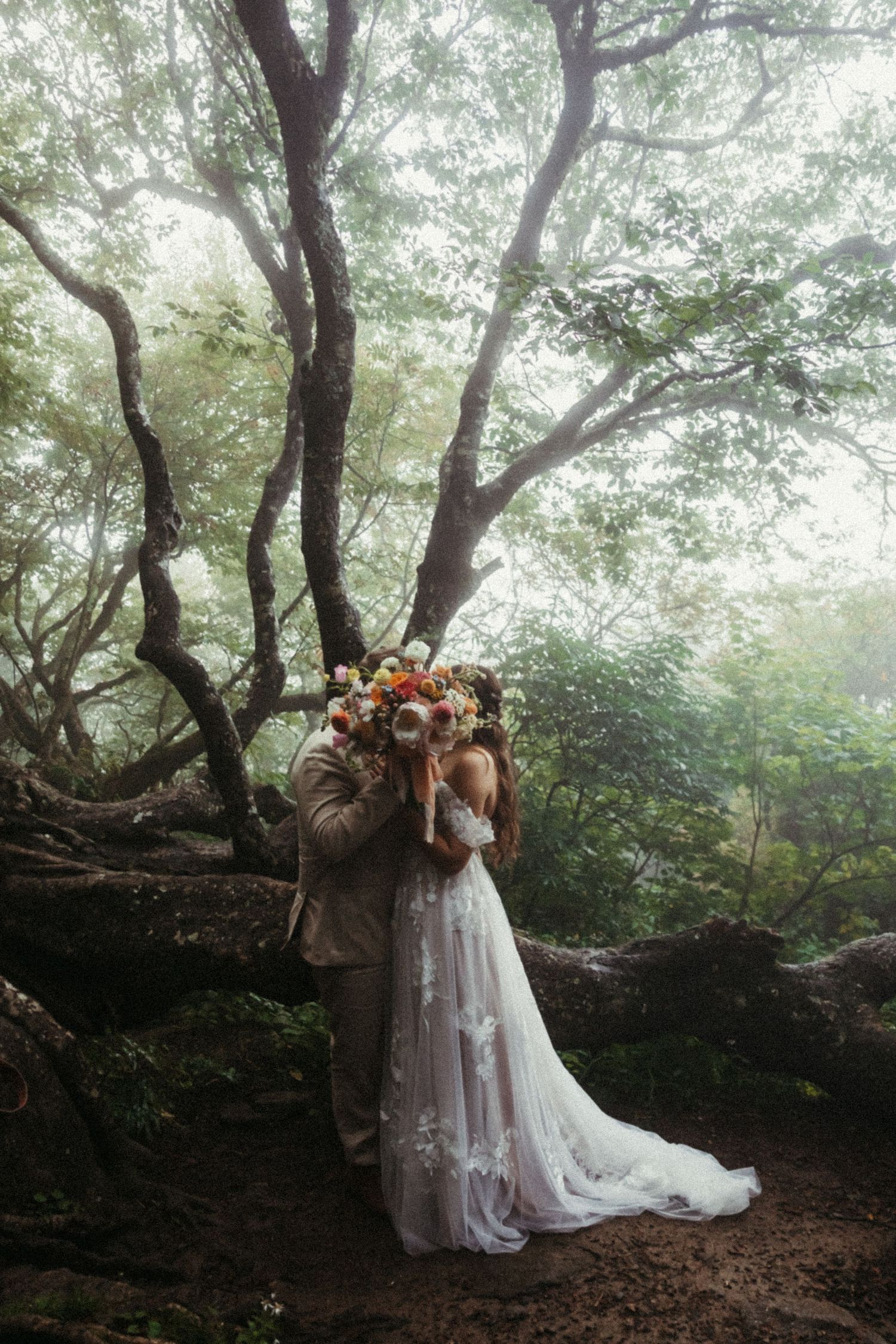 Bride and groom kissing behind a colorful bouquet in a misty forest, bride wearing a flowing white gown.