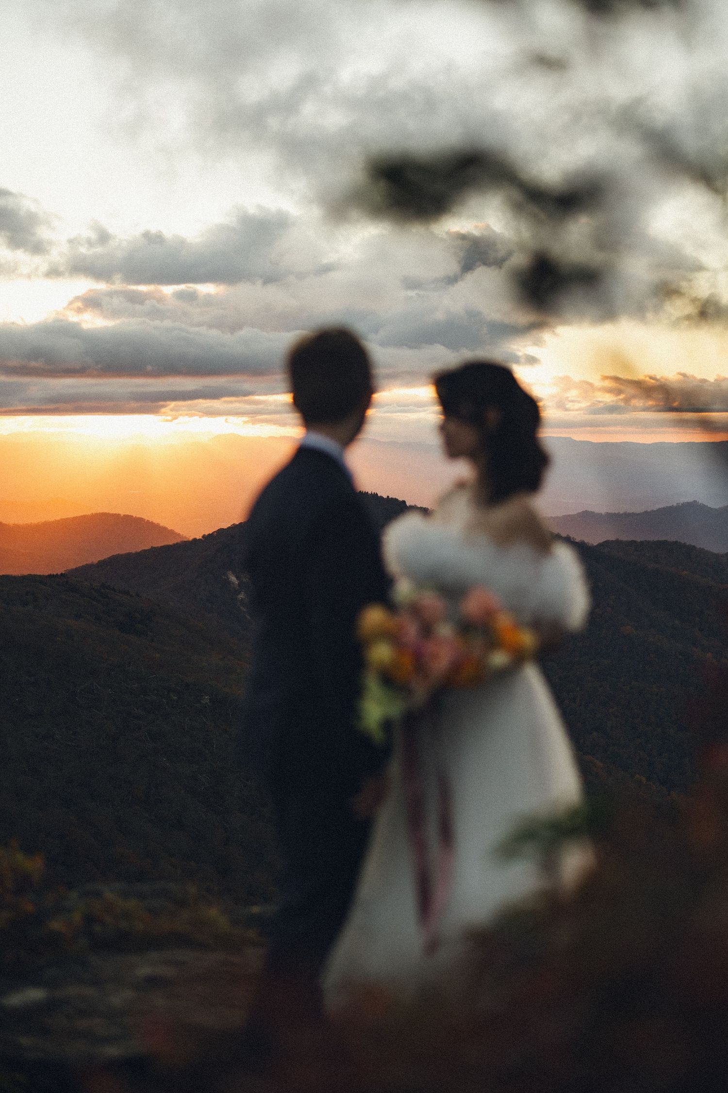 Silhouetted bride and groom standing on a mountain peak at golden sunset with vibrant orange sky backdrop.