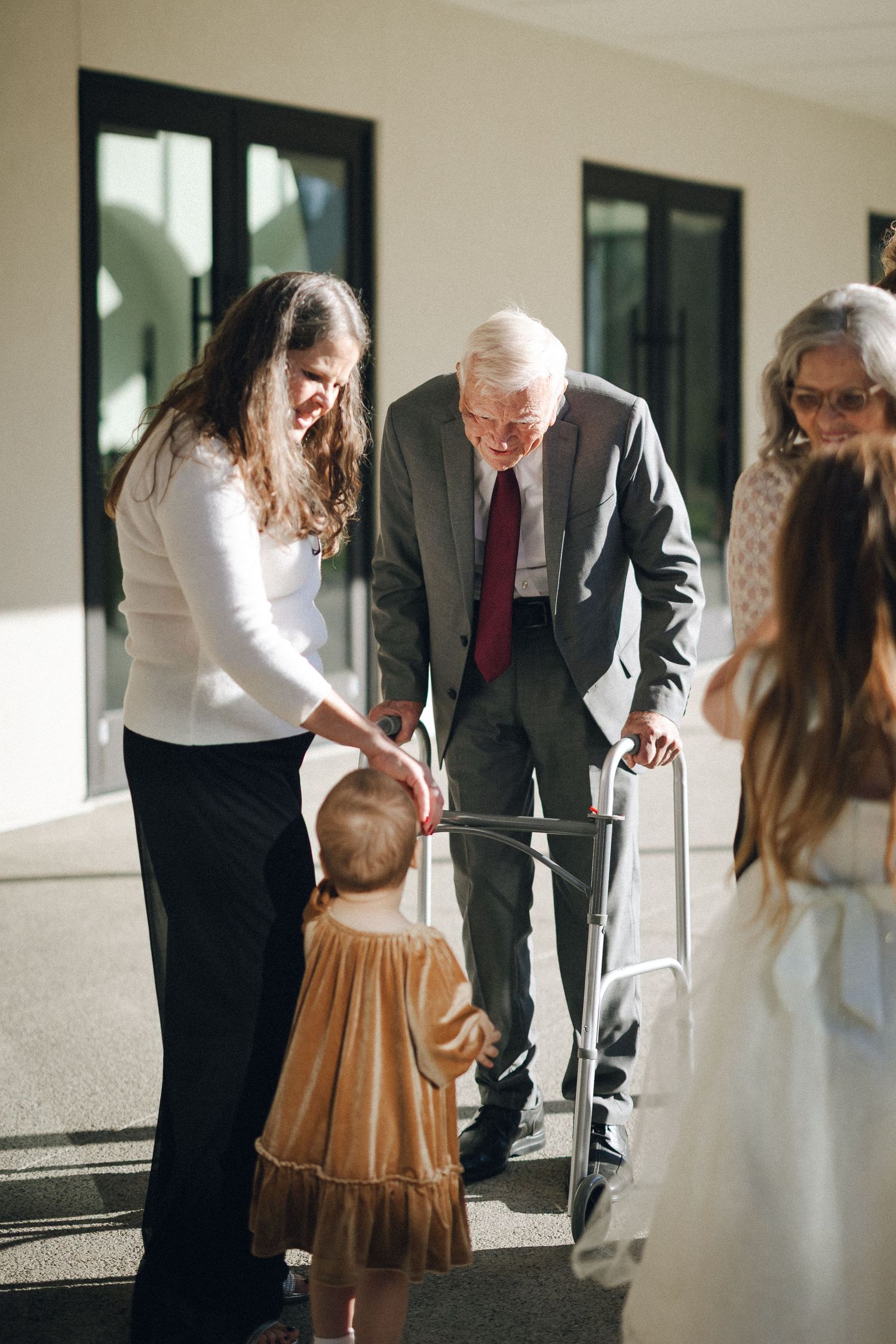 Elderly man with walker greeting toddler at wedding, assisted by woman in white top.