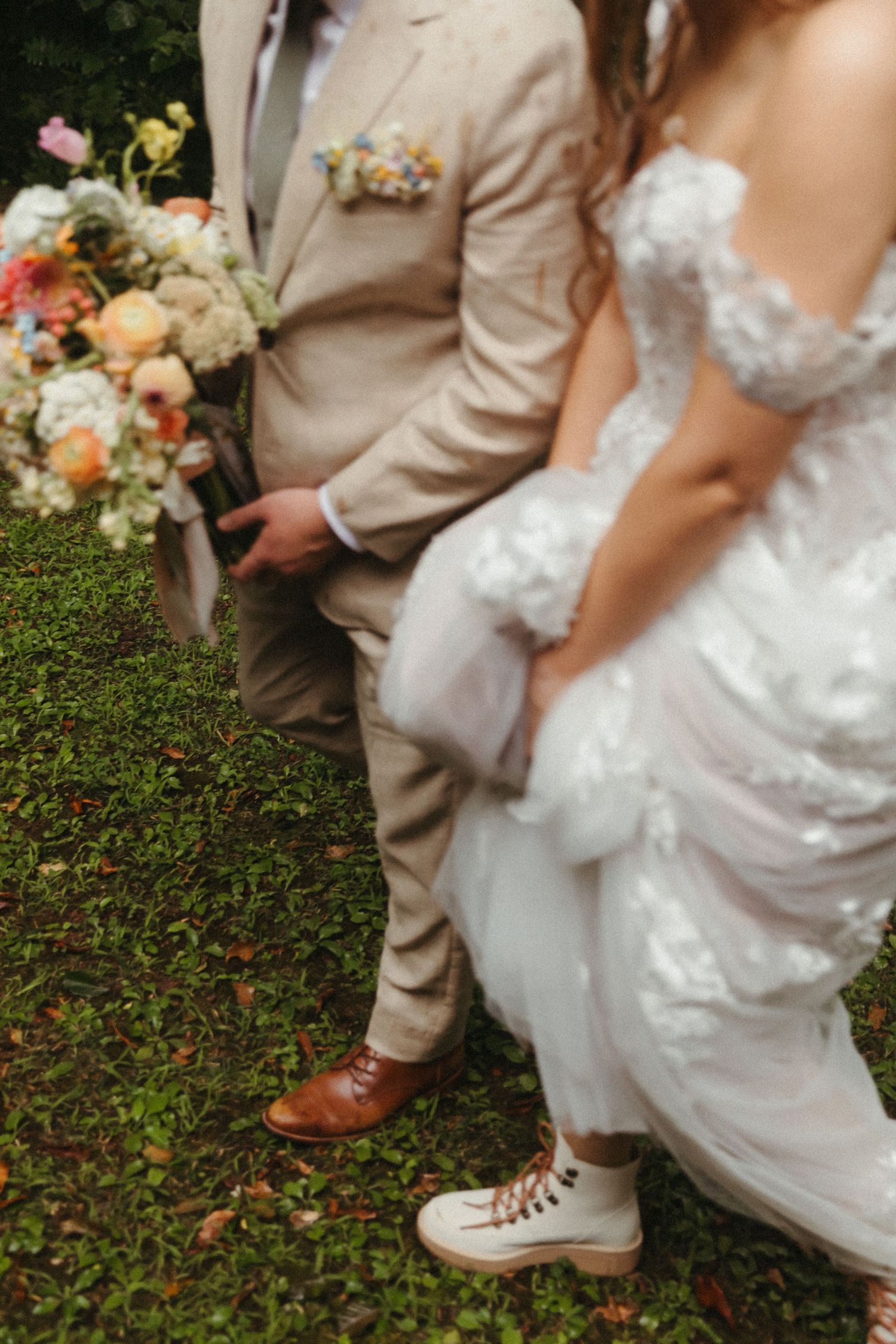 Bride in lace gown with hiking boots stands beside groom in tan suit on mossy ground.